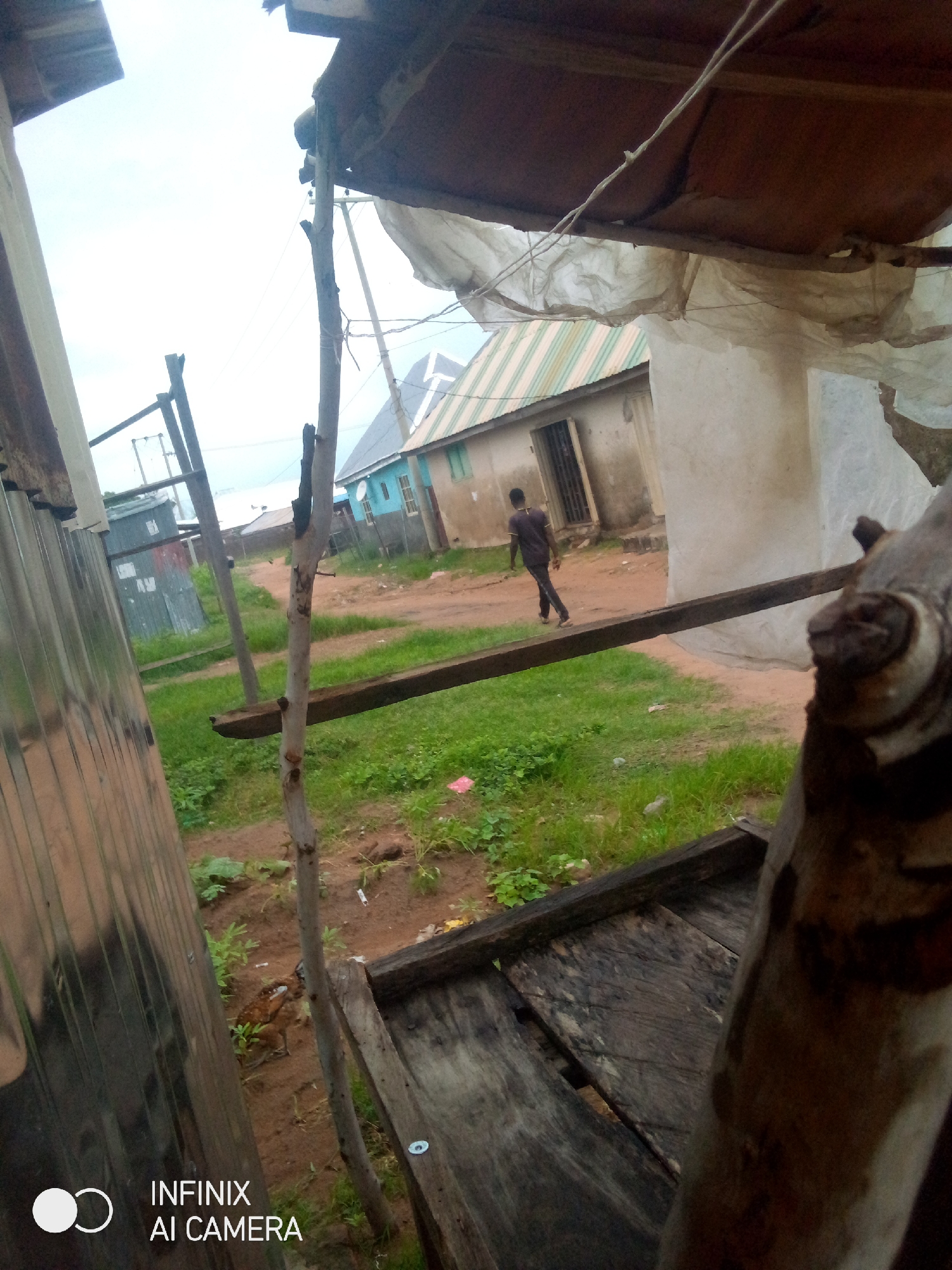 In the city of Jalingo, Nigeria, an outdoor scene unfolds with a dirt path winding through a grassy area. A solitary figure, a young man wearing a dark t-shirt and pants, walks away from the camera towards a cluster of modest buildings. One building features a light blue and green striped metal roof, while another is a light brown color with a door covered by a metal grate. Adjacent to these structures is a smaller, corrugated metal building. Overhead, a makeshift shelter is constructed with wooden poles and a brown, weathered roof, draped with translucent plastic sheeting. In the foreground, a rough, wooden structure, possibly a bench or table, occupies the lower right quadrant. A thin, vertical wooden pole stands in the mid-ground, supporting a horizontal wooden beam. The sky is overcast.