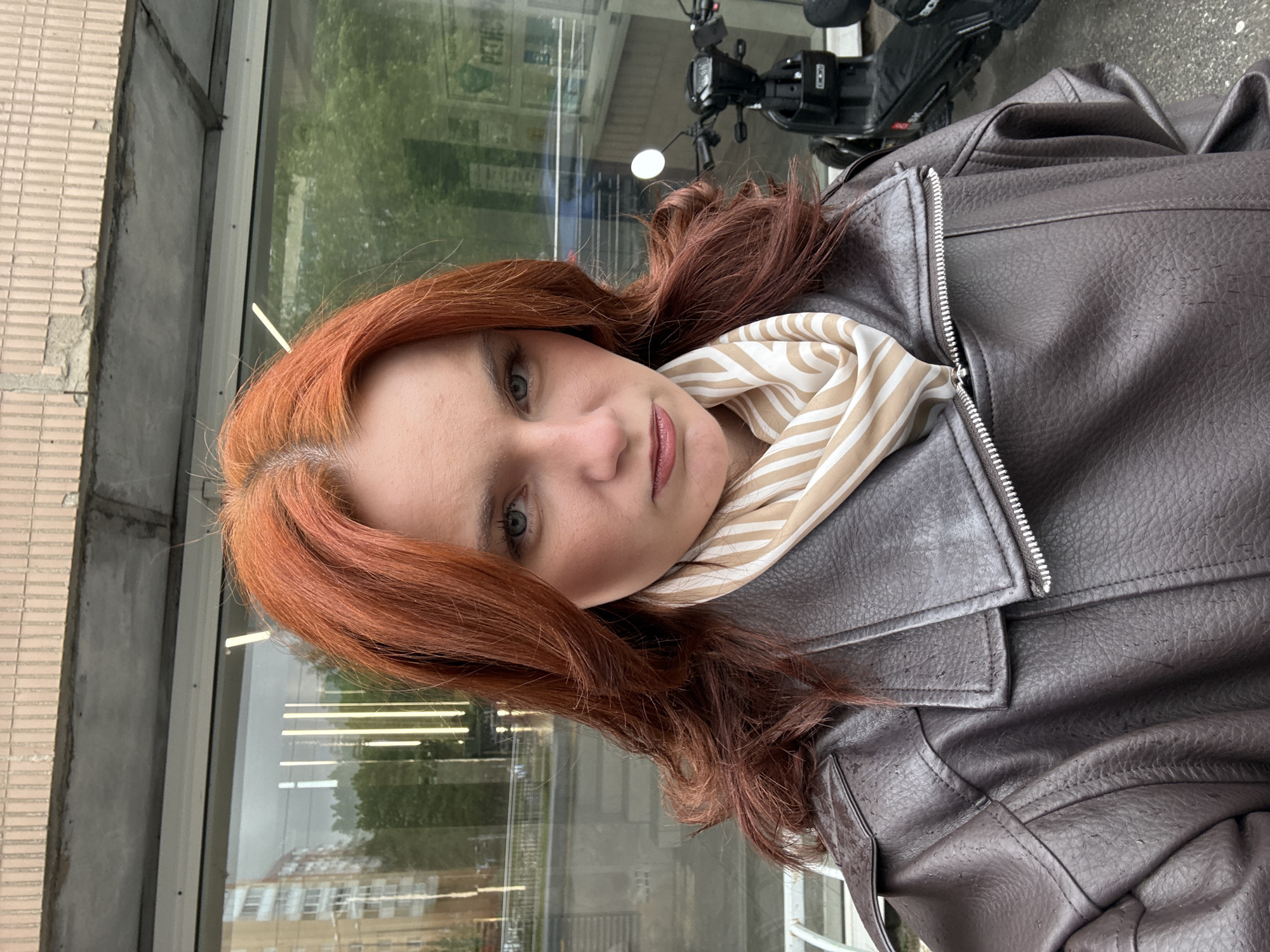 A woman with red hair and fair skin wears a brown leather jacket and a beige and white striped scarf. She stands outside a building with a glass wall reflecting the surrounding environment. A parked moped is partially visible in the background, as is the cityscape of Saint Petersburg, Russia, reflected in the glass. The overall setting suggests a casual, urban environment.