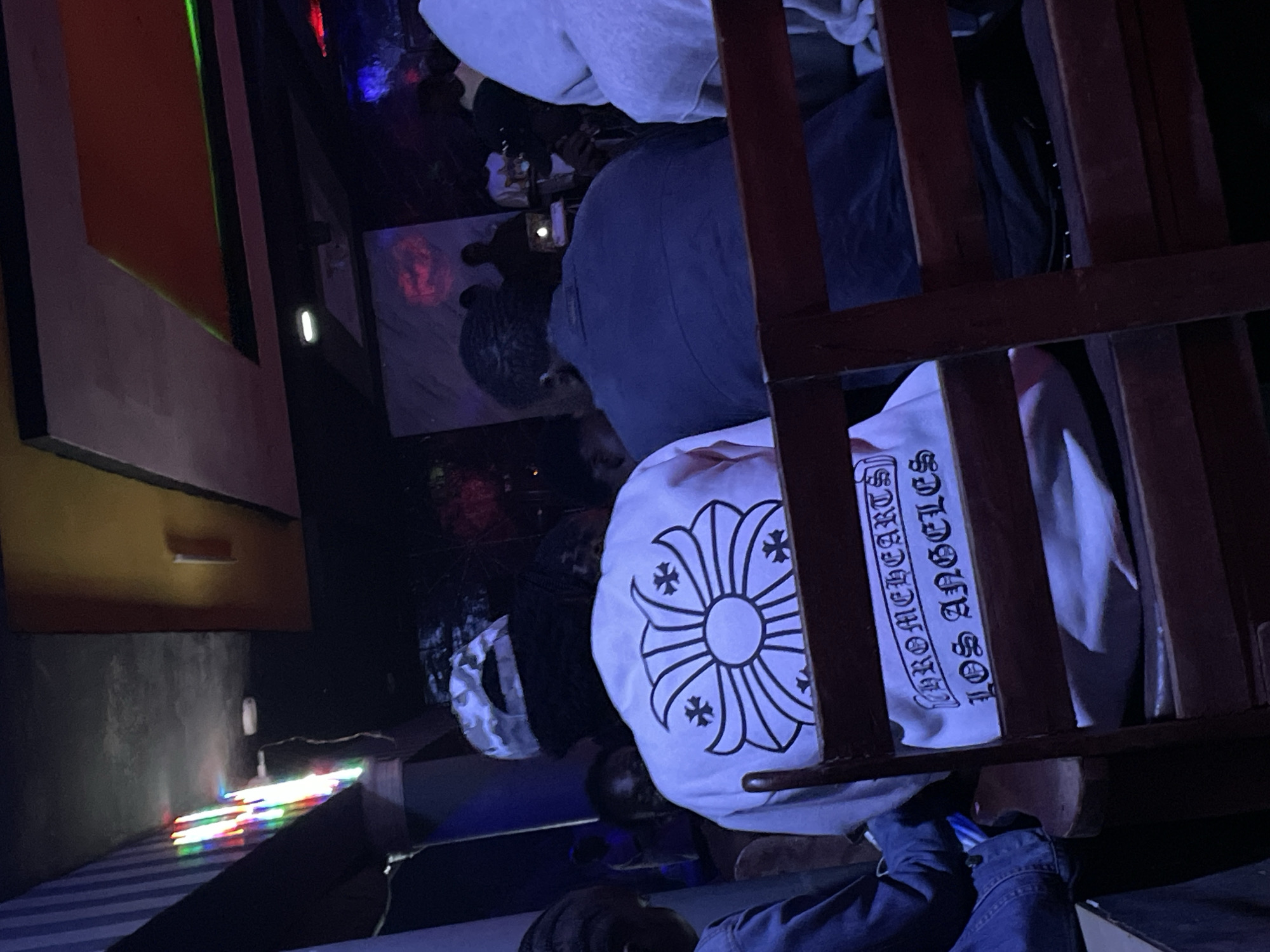 The image shows a dimly lit indoor scene, likely a bar or club, with multiple people visible. In the foreground, a person wearing a white t-shirt with a black graphic design of a flower-like emblem and the text "CHROMEHEARTS LOS ANGELES" is seated. To the right, another person in a dark blue shirt is also seated. In the background, more individuals are present, and a DJ booth with equipment is visible. Colorful lights are illuminating the space from above and on the left side of the image, creating a vibrant atmosphere. The overall setting suggests a social gathering or entertainment venue.
