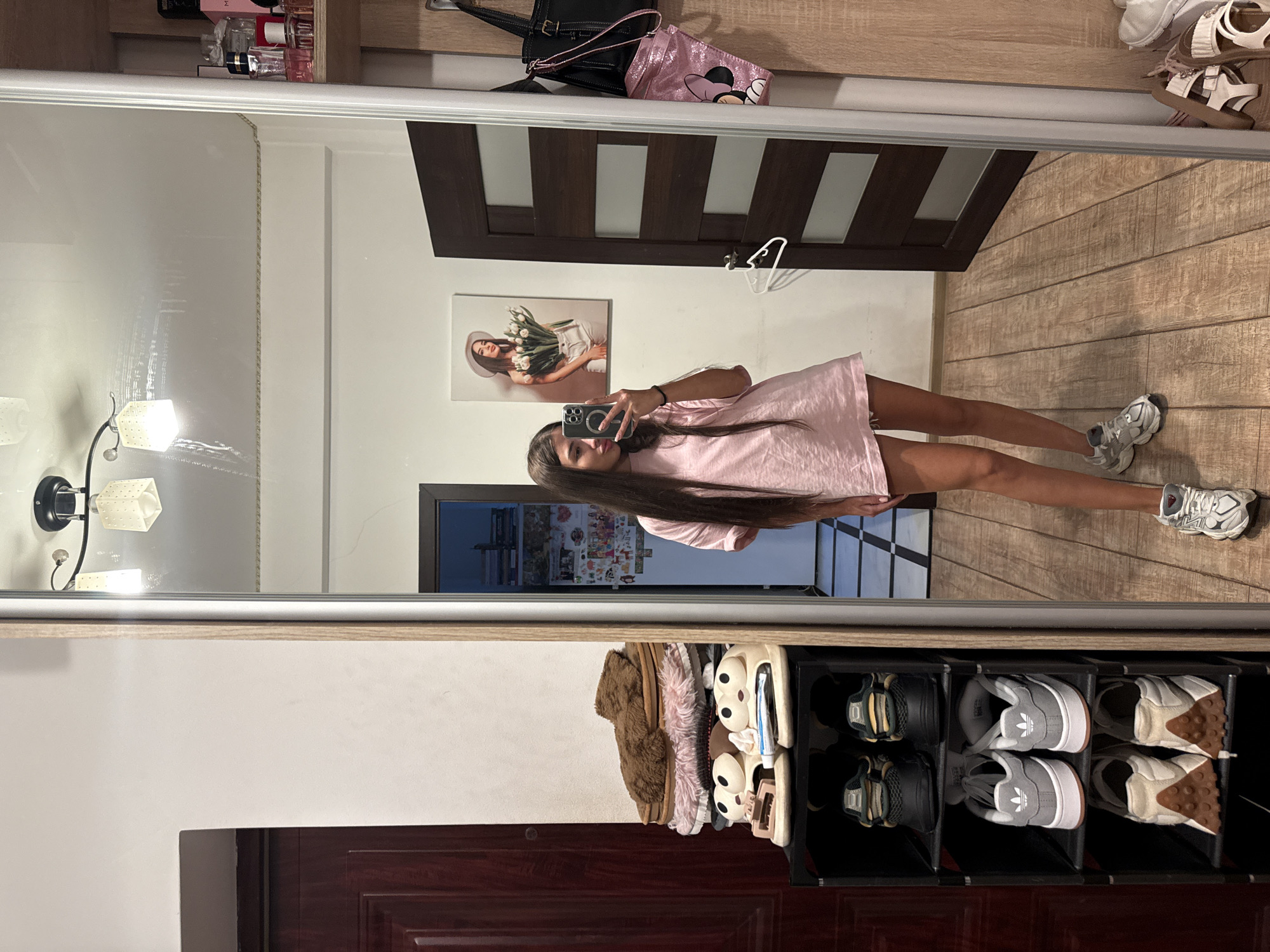 The image depicts an indoor scene, likely a hallway or dressing area, showing a young woman taking a mirror selfie. She is wearing a pink oversized t-shirt and shorts, and white athletic shoes. Her long dark hair is braided. She is standing in front of a large mirrored closet door.
To her left, a dark brown wooden door is visible, with a wooden frame. Next to the door is a tall, black, multi-tiered shoe rack containing several pairs of shoes and slippers. On top of the shoe rack are fluffy brown and pink slippers. Below them are several pairs of sneakers.
The mirrored closet door reflects the room behind the woman. A wall with a framed picture of a woman holding tulips is visible on the left side of the reflection. A dark brown interior door with horizontal glass panels is seen further back in the reflection. A black handbag and a pink bag with a Minnie Mouse design are hanging on the wall to the right of the mirrored closet door. Perfume bottles are displayed on a shelf above the hanging bags. The floor is made of wooden planks. The ceiling has a modern light fixture. The scene appears to be in a residential setting. No specific landmarks or indicators of Chișinău, Moldova, are present in the image.