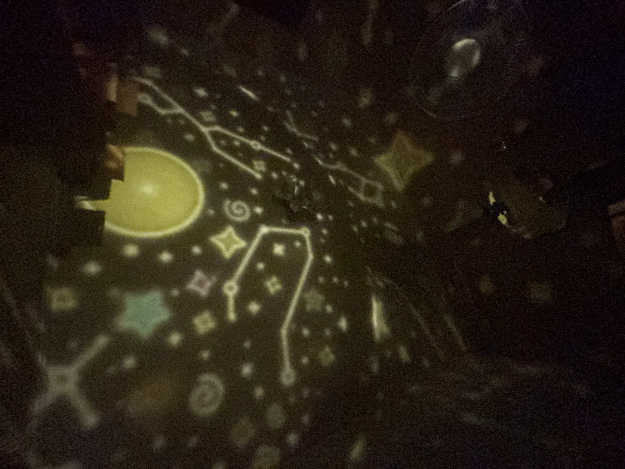 The image displays a dark room illuminated by a star projector. The projector casts luminous, stylized shapes of stars, constellations, and geometric patterns onto the ceiling and possibly walls. Some of the visible shapes include a large yellow circle, a green star, and various lines connecting dots, forming constellation-like patterns. The overall impression is of a playful or decorative lighting effect, likely intended for children or ambiance. There is no discernible location in Kryvyi Rih, Ukraine, or any specific environmental context beyond a darkened interior space. No people or specific objects are clearly identifiable within the scene other than the projected light patterns.