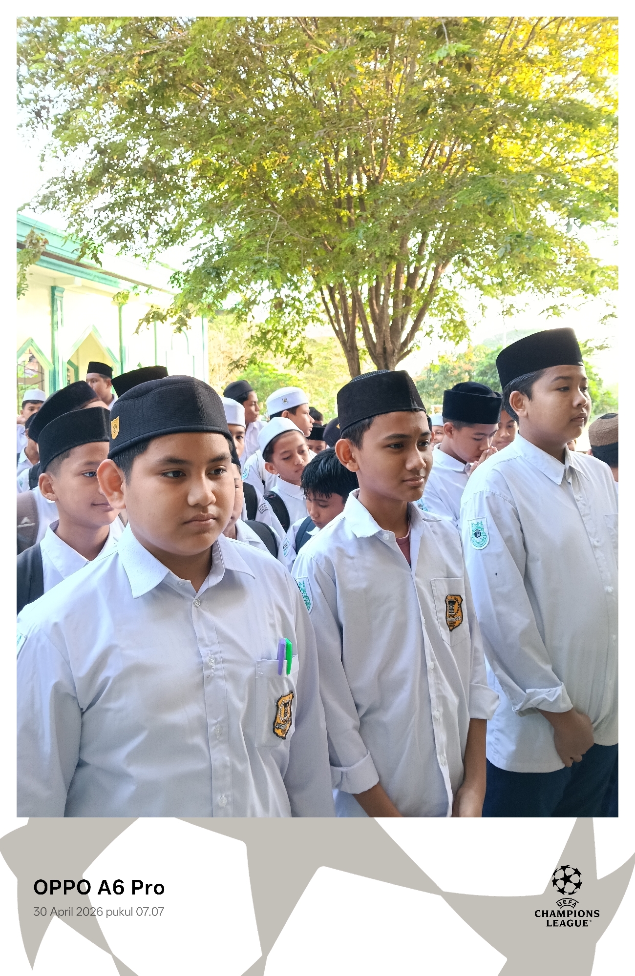 A medium shot depicts a group of boys, presumed to be students due to their matching attire, situated outdoors under a tree. They are dressed in light-colored, button-down shirts, some with sleeves rolled up, and dark-colored caps. Emblems adorn the shirts' pockets. The location, Lhokseumawe, Indonesia, is deduced from the image metadata, though specific contextual details are limited. The boys are standing in formation, possibly in a line or queue. Building elements with a similar color scheme can be seen at the background, which may indicate the location. The light suggests the photograph was taken during daylight.