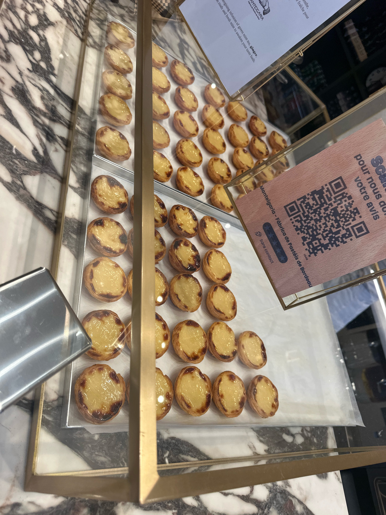 The image depicts a display case containing numerous Portuguese custard tarts, known as pastéis de nata. These tarts are arranged in rows on white paper within silver trays, which are themselves placed on a white surface with a marble-patterned backdrop. The display case has gold-colored framing and a glass front, through which the tarts are visible.
In the upper left quadrant of the image, a portion of a paper sign with text in French is visible, mentioning "mint tea" and "ginger." To the right of this, another sign with a QR code and text in French, "Mantelgaría - Fábrica de Pastéis de Bordeaux," is displayed. This sign also includes a star rating and the words "Sans contact." The presence of "Bordeaux" in the text on the sign strongly suggests that this establishment is located in Bordeaux, France.
The lighting in the scene is bright, illuminating the tarts and creating reflections on their glazed surfaces. The overall impression is one of a clean and well-presented confectionery display. No people are visible in the image.