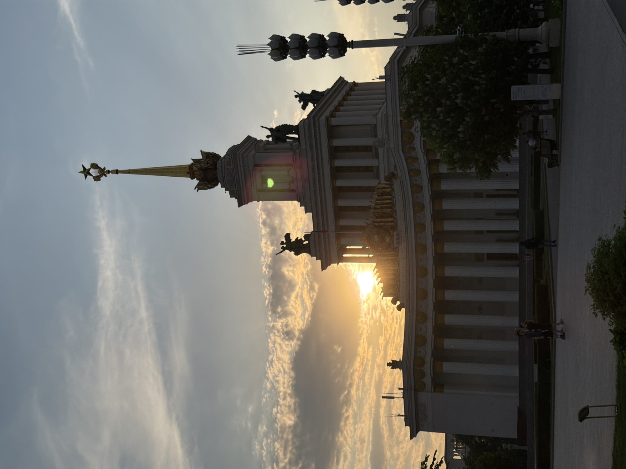 The image shows the Central Pavilion at the Exhibition of Achievements of National Economy (VDNKh) in Moscow, Russia, against a backdrop of sunset and clouds. The pavilion's architecture is visible, including its spire topped with a star. Figures and statuary adorn the building's exterior. People are walking on the path next to the building. The light is bright due to the sun setting. There are some trees surrounding the building as well as street lamps.