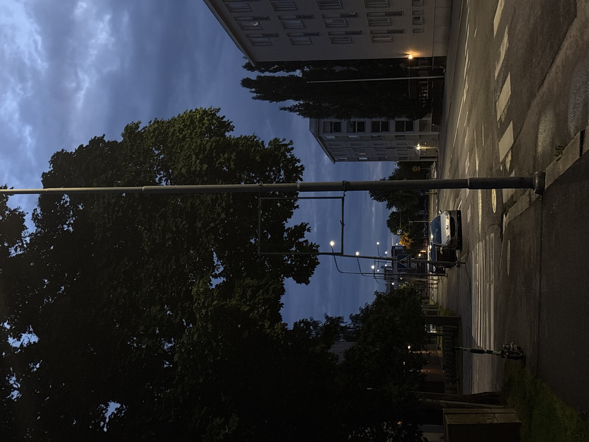 The image depicts a street scene in Pori, Finland, during twilight. On the left side of the image, a large tree with dark green foliage dominates the foreground, casting shadows. A grey lamppost stands tall in front of the tree. In the middle ground, a pedestrian crossing with white stripes is visible across a paved road. A parked electric scooter with black and white markings stands on the sidewalk near the crossing. Several cars are parked along the side of the road, and one silver car is crossing the pedestrian crossing. The road itself appears wet in places, reflecting the dim light.
On the right side of the image, two multi-story apartment buildings are visible. The building in the immediate foreground is light grey and has numerous windows. The building further back is also light grey with a similar window pattern. Between the buildings, tall, dark, slender trees stand out against the evening sky. The sky is filled with dark, cloudy formations, with hints of blue peeking through. Streetlights are visible along the road, some illuminated, casting pools of light. A pedestrian crossing sign with a blue background and a white silhouette of a pedestrian is mounted on a lamppost. A speed limit sign indicating "40" is also visible on a post. The overall atmosphere is calm and quiet, characteristic of an urban environment at dusk.