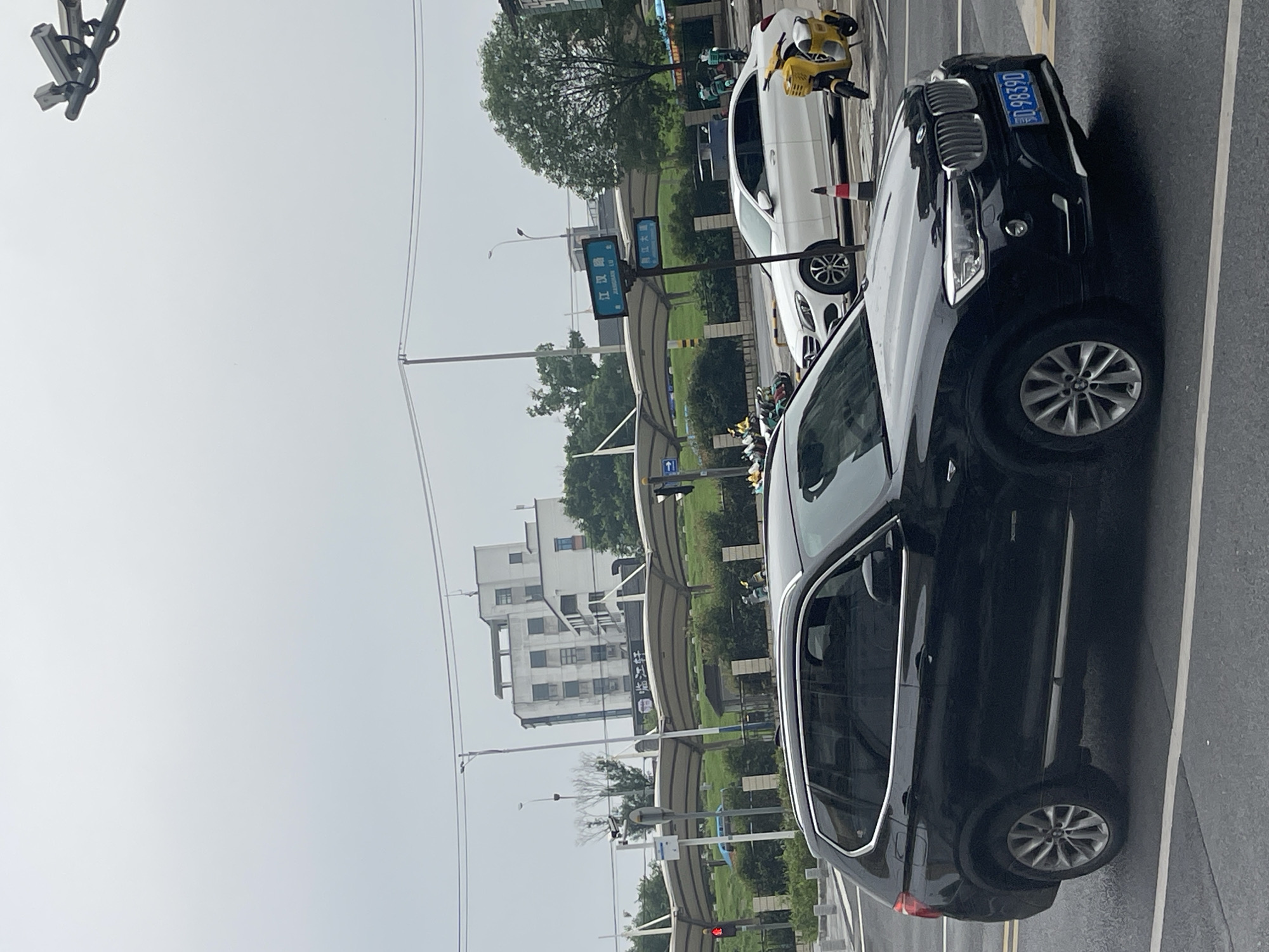 The image depicts an urban street scene at the intersection of Jianghan Road (江汉路) and Yanjiang Avenue (沿江大道) in Wuhan, Hubei Province, China, under an overcast sky. The road surface appears dark, possibly damp.
In the foreground, a black BMW SUV, bearing the Chinese license plate 鄂 D.9839D, is positioned on the roadway, facing slightly right. The vehicle has silver trim accents and a chrome front grille. To the right of the BMW, a white Mercedes-Benz sedan is partially visible, either parked or stopped, with an orange and white traffic cone positioned nearby. Adjacent to the Mercedes, a yellow electric scooter with a front basket is parked.
A street sign pole centrally located displays two blue signs: the upper sign reads "江汉路" (Jianghan Road) and the lower sign reads "沿江大道" (Yanjiang Avenue), confirming the intersection. Behind the street signs, a series of arching, light-colored canopy structures span a green landscaped area with bushes. Multiple shared electric scooters, in both yellow and green liveries, are parked on a paved section behind these canopies. A red traffic light is visible on the left side of the frame.
In the background, a multi-story white building with multiple windows and architectural details is discernible. Various trees and green foliage are scattered throughout the scene. Overhead utility wires traverse the sky, supported by utility poles. In the upper right corner, two surveillance cameras are mounted on a pole, directed towards the road below. The sky is uniformly light gray and overcast.