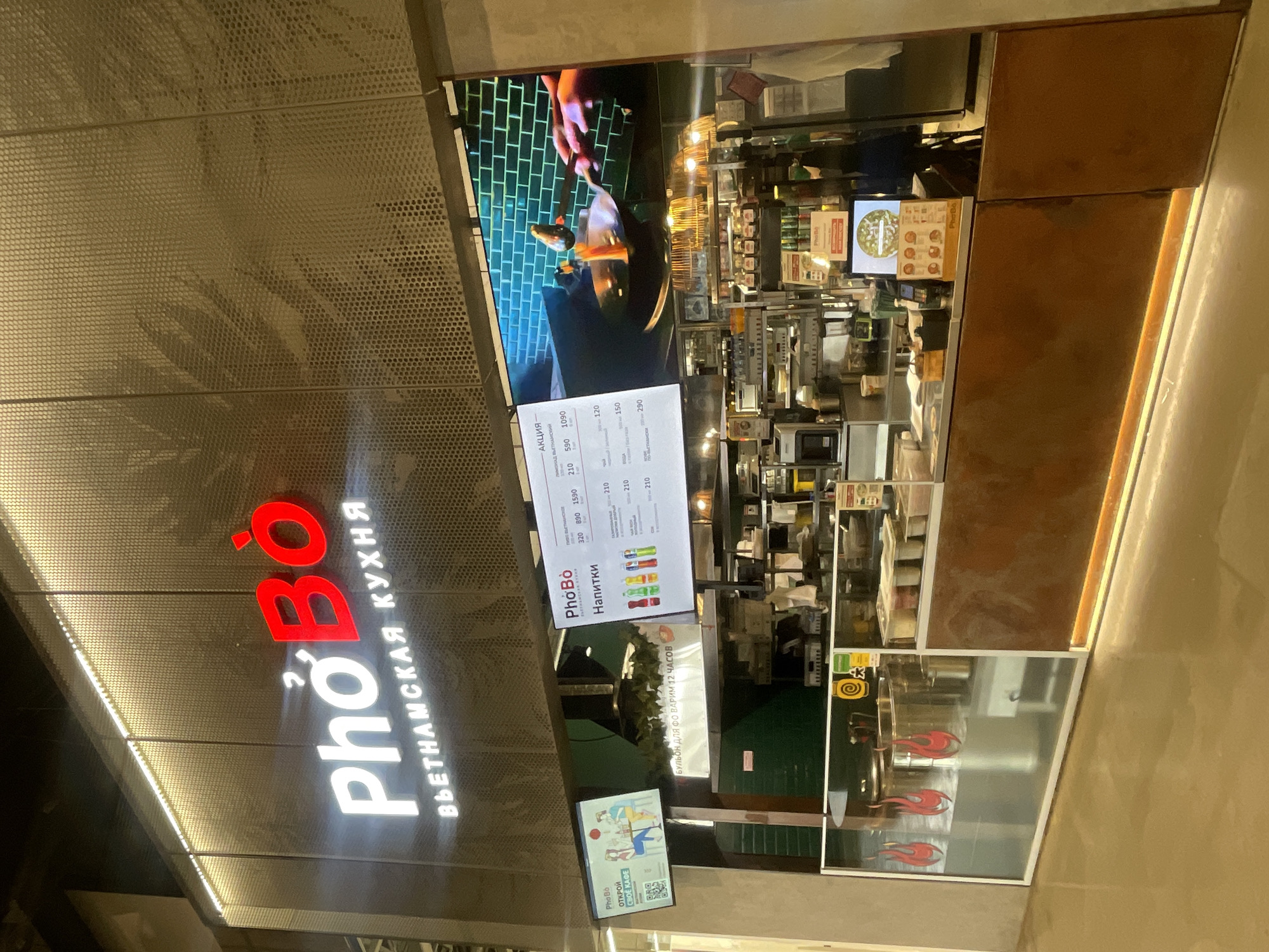 Here is a detailed description of the image:
**Scene:** The image shows the exterior of a "Pho Bó" restaurant, offering Vietnamese cuisine. The façade of the restaurant is a perforated metal panel, where the name "Pho Bó" is displayed in white and red letters, with "ВЬЕТНАМСКАЯ КУХНЯ" (Vietnamese Cuisine) written beside it in Cyrillic. The restaurant appears to be situated in an urban area.
**Signage:** The restaurant features a menu sign showcasing offerings and prices in Russian. There is also promotional material visible, including a sign mentioning that the broth is cooked for 12 hours, and another promoting franchise opportunities.
**Environment:** The environment suggests an interior view of the restaurant's cooking area and preparation area. A video screen shows food being prepared. The overall setting has modern design elements, with a combination of metallic and possibly wooden finishes.
**People:** People are visible inside the restaurant. However, their roles are not clearly discernible.
**Objects:** Key objects visible are the restaurant's signage, the menu, cooking equipment, food preparation items, and various promotional displays.
**Actions:** The actions within the image are mainly related to the operation of the restaurant.
**Location:** This appears to be located in the city of Krasnoyarsk, Russia.