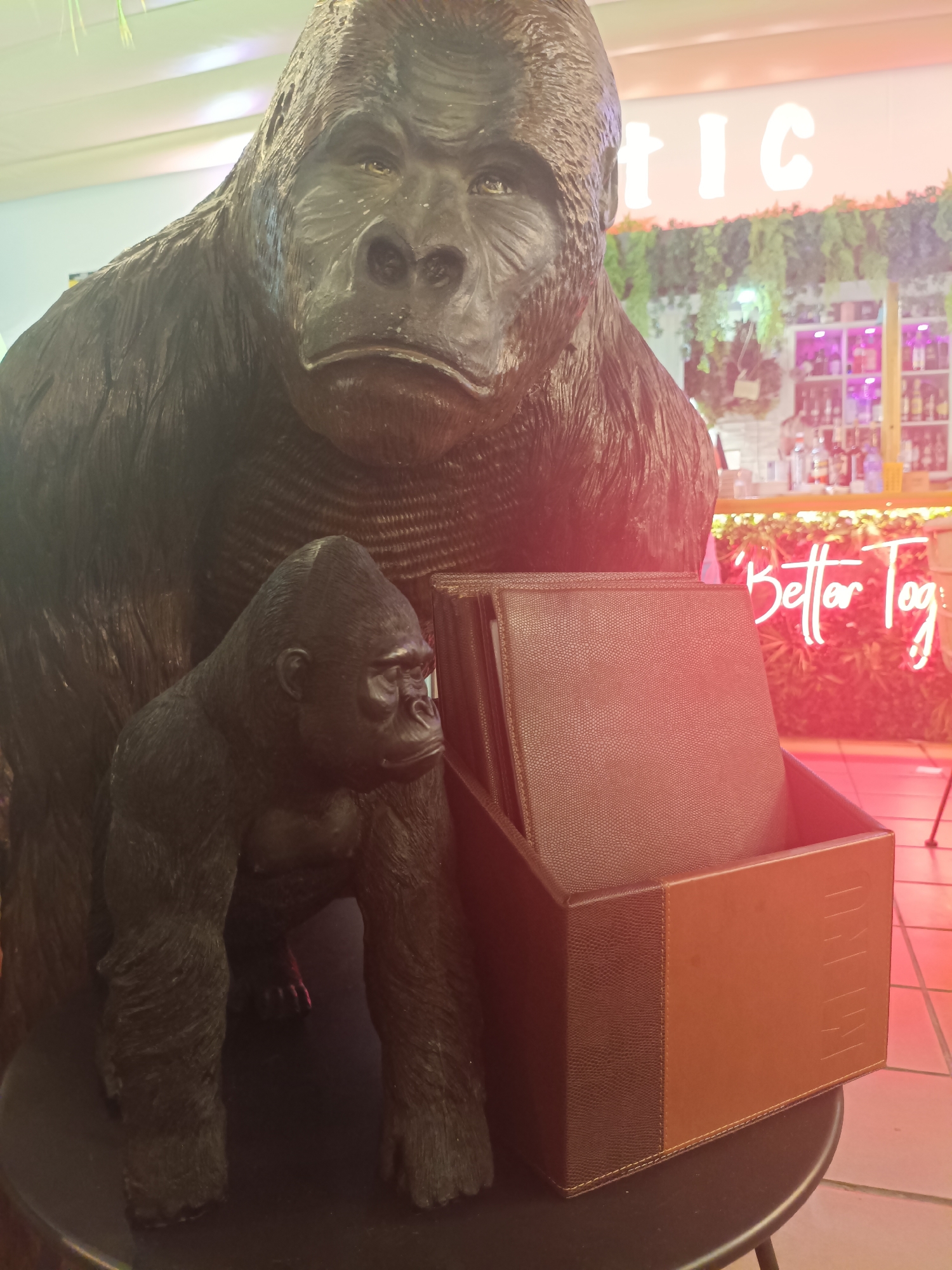 The image shows a close-up view of a life-sized gorilla statue and a smaller gorilla statue in front of it, both appearing to be made of dark, textured material. In front of the gorillas is a brown leather desk organizer containing several menu-like folders. Behind the statues, a neon sign reads "Better Tog" in pink lettering, set against a backdrop of greenery and a bar area with bottles visible on shelves. The scene appears to be indoors, possibly a restaurant or themed establishment. The location is identified as San Gabriel, Spain.