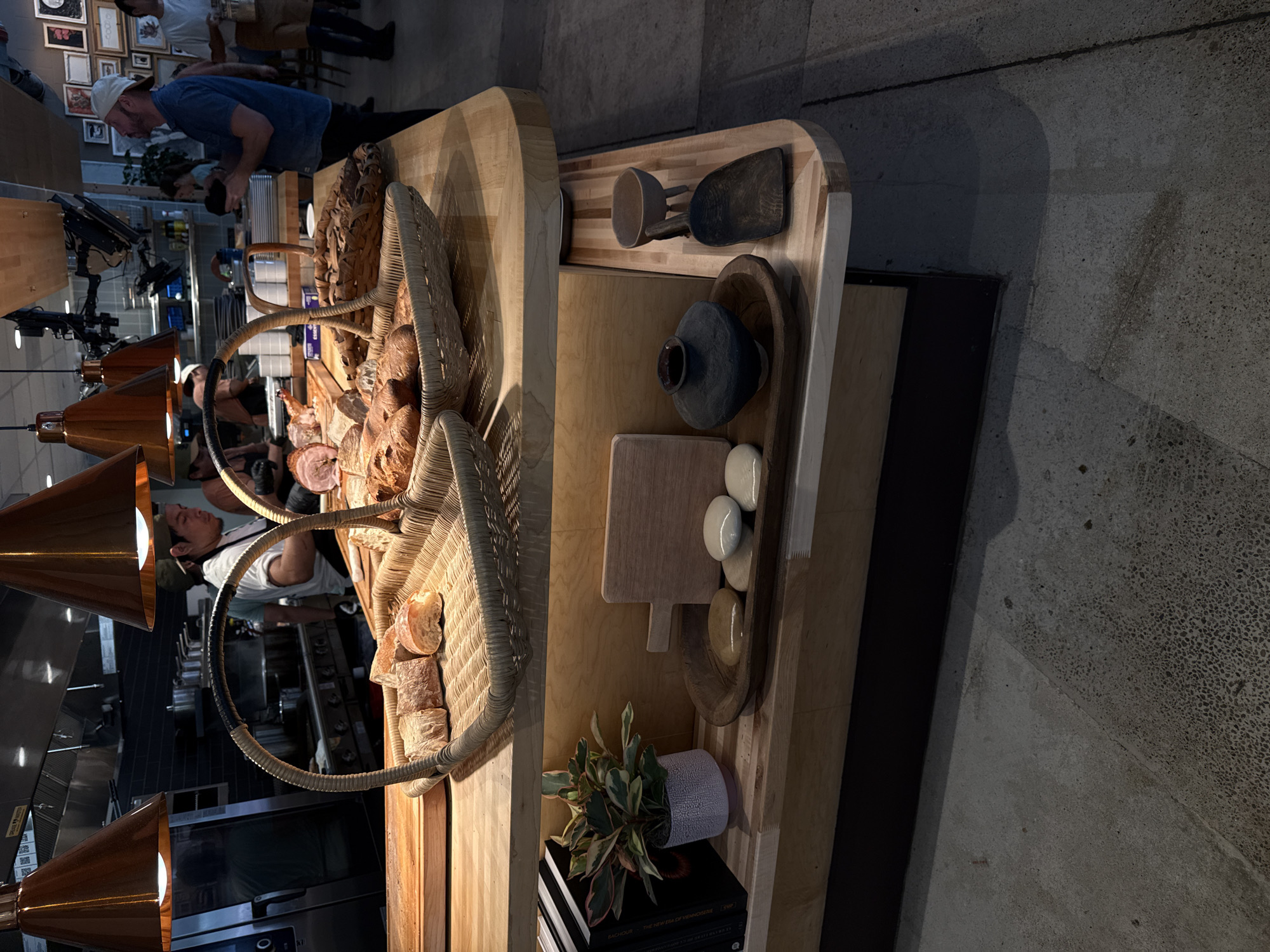 The image shows a bakery setup, likely a counter, with various food items displayed in baskets and on wooden boards. Several individuals are visible, including a man in a blue shirt near the counter and other people in the background, possibly employees or customers. The setting appears to be inside a restaurant or bakery.
Specifically, the image depicts goods sold at The Swedish Hill Bakery in Austin, Texas, USA.