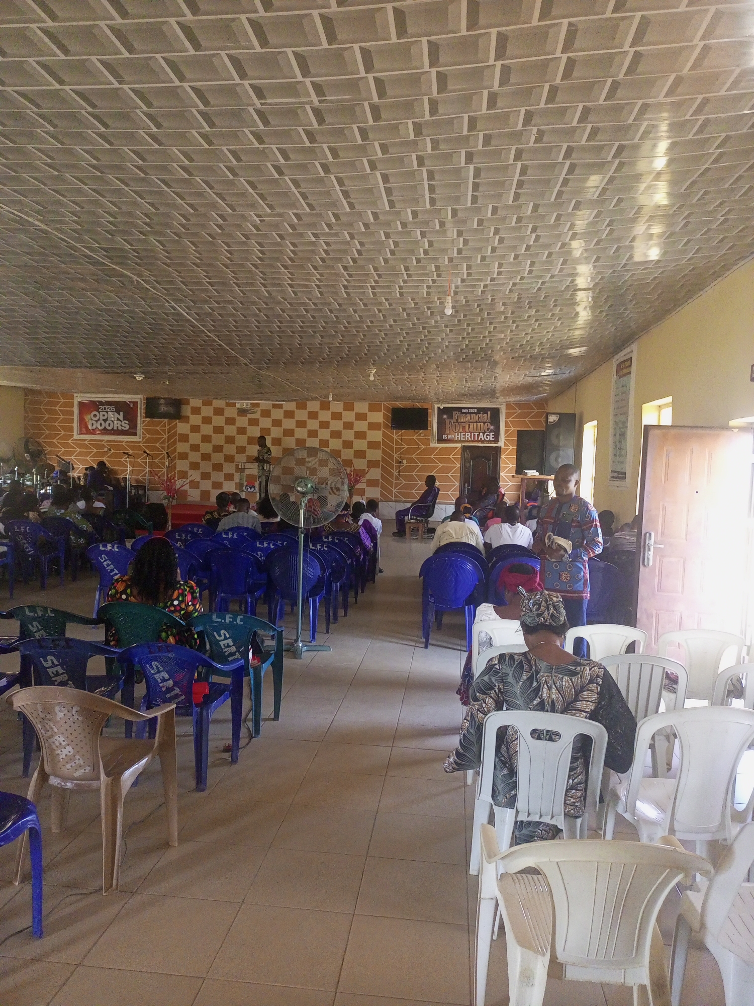 The image depicts an indoor gathering, likely a church service, in Serti, Nigeria. The scene is characterized by rows of blue and white plastic chairs arranged on a tiled floor. The walls are decorated with a checkered pattern in shades of orange and brown, and a banner reading "2026 OPEN DOORS" is visible on the left wall. Another banner on the right wall displays text that appears to be related to "FINANCIAL FORTUNE IS MY HERITAGE". Several individuals are seated in the chairs, facing towards the front of the room where a podium is visible. A man in traditional attire is standing near the podium, possibly addressing the congregation. A large standing fan is positioned in the middle of the room. The ceiling is a metallic grid pattern. The overall atmosphere suggests a community event or religious service.