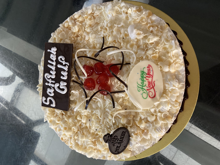 A round celebratory cake is centrally positioned on a golden, corrugated cardboard base, resting on a reflective, dark gray table surface. The cake's upper surface is densely covered with numerous small, irregularly shaped, light-colored flakes, consistent with white chocolate shavings or almond slivers. Dark chocolate frosting or drips are visible along the bottom circumference of the cake where it meets the golden board.
Key decorations include:
1. A rectangular dark chocolate plaque positioned at the upper-center, displaying the text "Saifullah Gulf" in white icing.
2. A cluster of approximately seven glossy red glacé cherries or maraschino cherries near the cake's center, surrounded by thin, curled strands of dark and white chocolate.
3. A small, oval-shaped dark chocolate plaque located on the left side, embossed with "MR. BAKER" and a stylized chef's hat logo.
4. A larger, oval-shaped white chocolate plaque situated at the lower-center, featuring "Happy New Year" written in decorative green and red script.
The reflective table surface shows indistinct, shadowed reflections. No individuals are visible, and no specific actions or interactions are depicted. The cake is branded by "MR. BAKER," a bakery chain with locations in Dhaka, Bangladesh. However, the image itself provides no visual information to identify a specific location within the city of Dhaka.