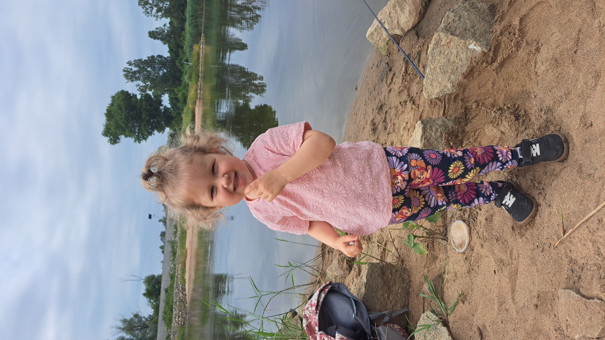 A young child, appearing to be a girl with blonde curly hair pulled into a top knot, stands on a sandy riverbank. She is wearing a pink textured t-shirt and patterned leggings featuring flowers and cartoon characters. The child is smiling and has her hand near her mouth. To her left, a backpack is visible. Behind her, a calm body of water reflects the overcast sky and trees lining the opposite bank. The environment suggests a natural setting, likely a park or recreational area near a river. There is no indication of the specific location within Krosno Odrzańskie, Poland.