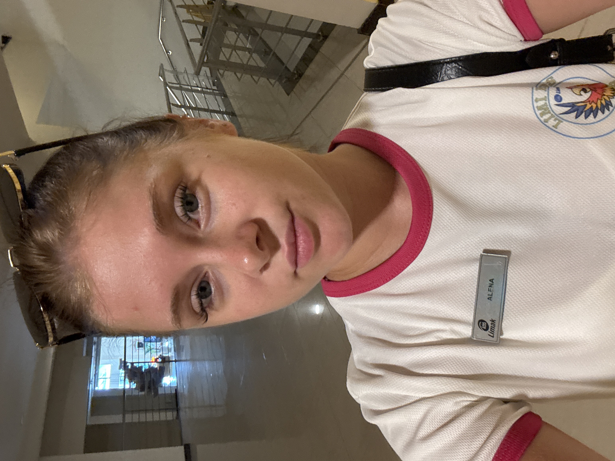 A young Caucasian woman with blonde hair, wearing a white shirt with pink trim and a black purse strap, is posing for a selfie indoors. She has on sunglasses pushed up on her head, and a name tag on her shirt reads "Limak ALENA". The background appears to be a hotel lobby or a similar establishment, with a tiled floor, railings, and stairs visible. The woman is the primary subject and is centrally framed in the shot. The setting, particularly the "Limak" branding on the name tag, suggests the location is likely a Limak hotel in Aksu, Turkey.