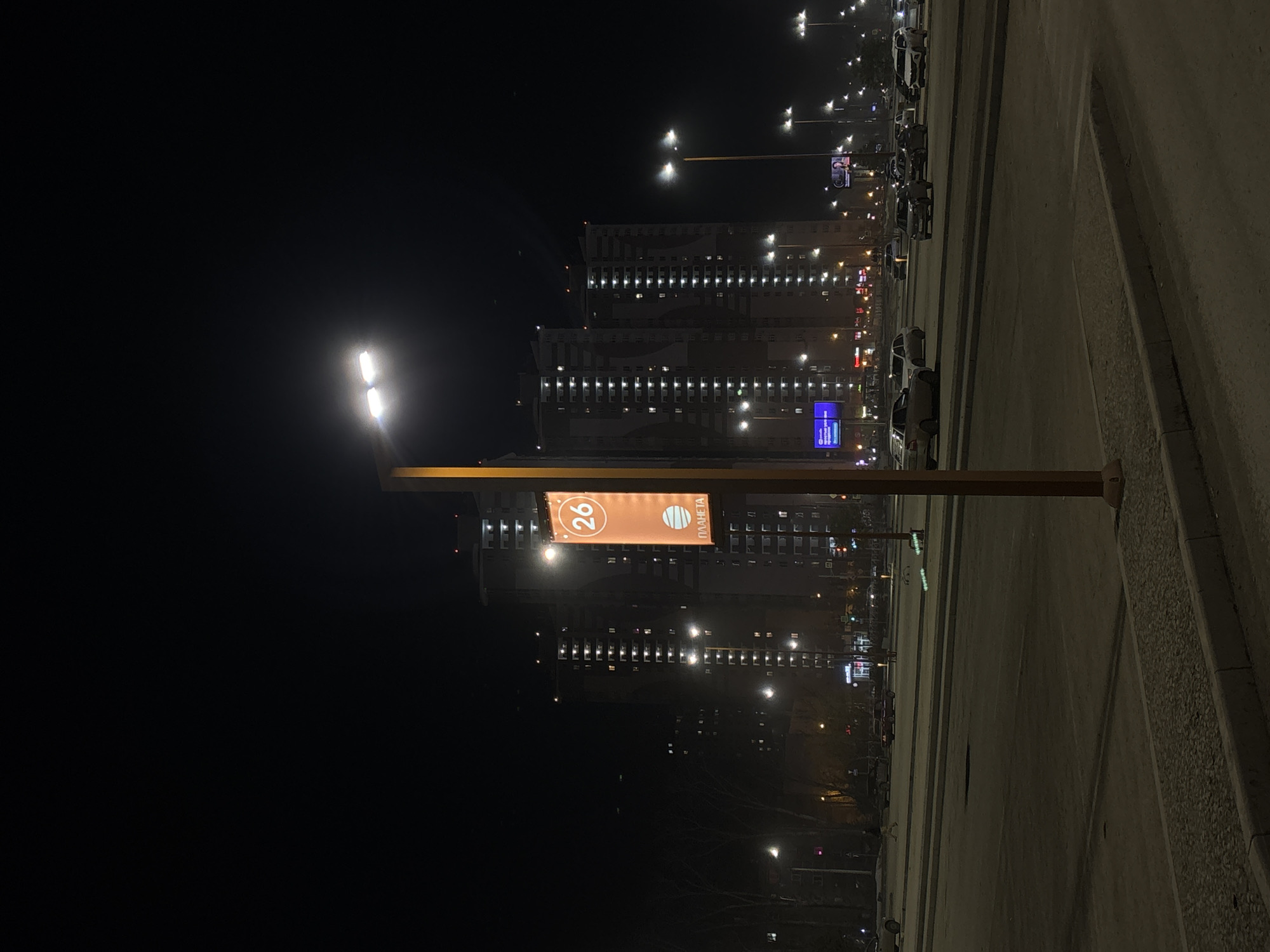 The image is a nighttime view, possibly from a window or high vantage point, overlooking a street in what appears to be the city of Perm, Russia. The buildings are tall and illuminated, showcasing multiple lit windows. The street has a number of cars parked alongside it. Streetlights are also present, providing illumination along the street and adjacent areas.
A prominent feature is a tall street sign or advertisement post with a sign that features the number 26 and the Russian word "ПЛАНЕТА" (Planeta). The sign is illuminated and appears to be the name of a complex. A lamppost is positioned directly in front of the buildings.