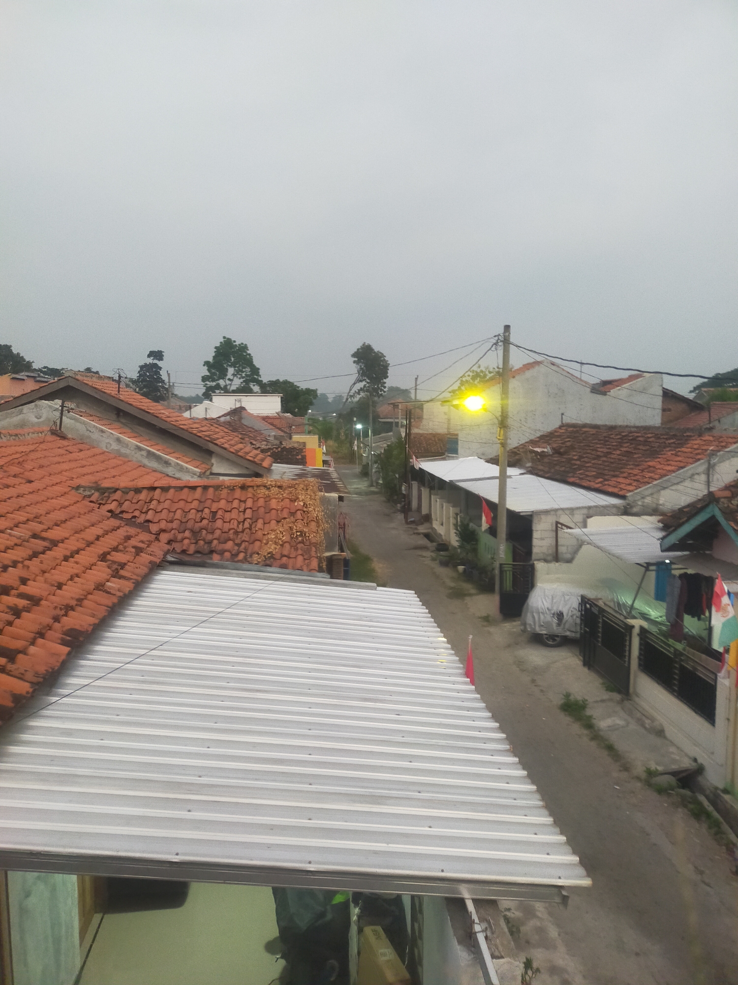 This is an aerial, eye-level, wide shot of a street in a residential area in Tegalandong, Indonesia. The scene is overcast, suggesting it is either early morning or late afternoon. In the foreground, a corrugated metal roof is visible, with a traditional terracotta-tiled roof extending to the right. The street itself is paved and appears to be a residential lane with houses on either side. A streetlamp with a bright yellow bulb is illuminated on the right side of the street. There are several houses with terracotta tile roofs, some with visible chimneys. Some houses have modern, flat, silver roofs. Several Indonesian flags are displayed, indicating a possible national holiday or event. A vehicle covered with a tarp is parked on the right side of the street, alongside a black metal fence. Laundry is hanging on a clothesline near the fence. Vegetation is sparse, with some trees visible in the distance and small plants growing along the side of the street. The overall impression is that of a quiet, densely populated residential neighborhood.