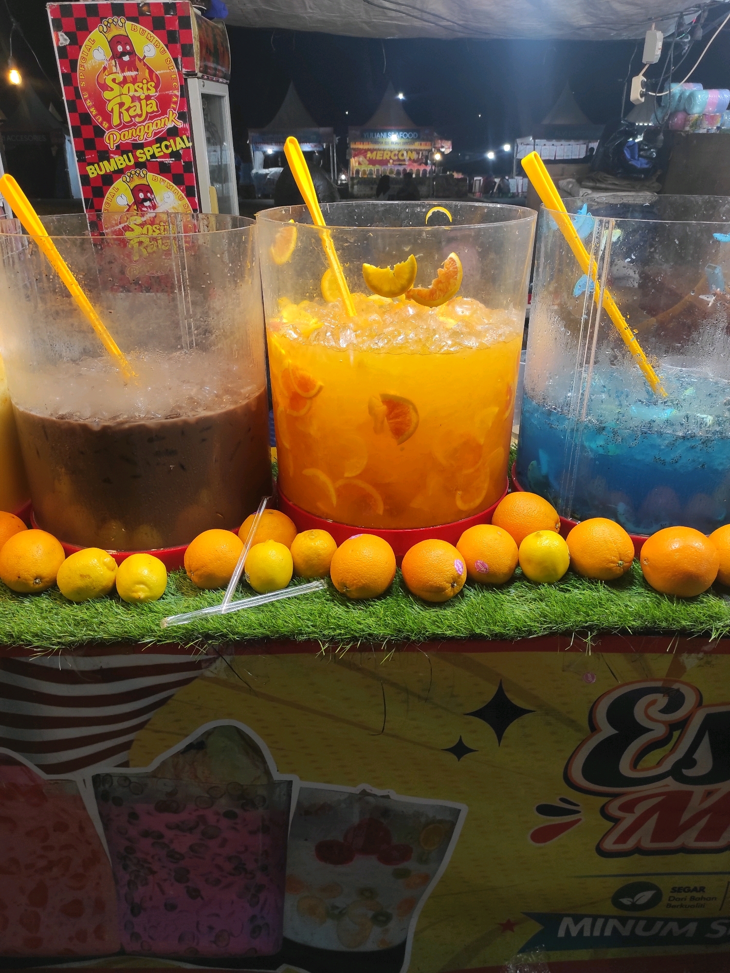 This image captures a street food stall in Central Jakarta, Indonesia, likely at night, as indicated by the artificial lighting. The stall displays an array of colorful drinks in large clear dispensers. The leftmost dispenser holds a dark, possibly chocolate-flavored beverage. Next to it is a large dispenser filled with a bright orange drink, garnished with slices of orange and ice. The rightmost dispenser contains a vibrant blue liquid, also with ice.
In front of the dispensers, arranged on a green artificial grass surface, are several whole fruits, including oranges and lemons, presumably for use in the drinks. Two yellow straws are visible, one in the orange drink and another in the blue drink. A sign for "Sosis Raja Panggank Bumbu Special" is prominently displayed behind the dispensers, suggesting a food item also sold at the stall. Further in the background, tents and other stalls are faintly visible, indicating a market or fair setting. The overall impression is of a lively nighttime street food scene.