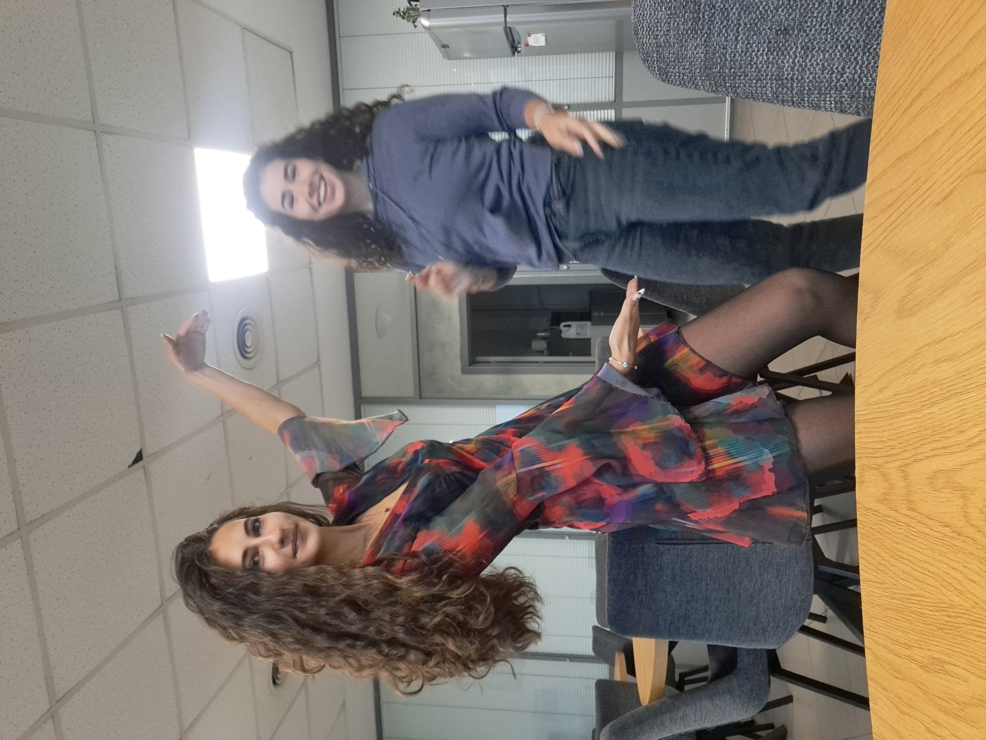 The image, oriented 90 degrees clockwise from its natural upright position, captures two women in a brightly lit indoor space, likely an office breakroom or lounge area in Sofia, Bulgaria.
In the foreground, a woman with long, curly dark brown hair is seated on a gray upholstered chair. She is wearing a long-sleeved top or dress featuring a vibrant, abstract pattern with shades of red, orange, purple, and blue. Her legs are crossed, clad in black sheer tights, and a delicate bracelet adorns her left wrist. She gazes towards the camera with a gentle smile, her right arm raised in a posed gesture.
Behind and slightly to her left, a second woman with long, curly dark brown hair stands, dressed in a dark blue long-sleeved top and dark jeans. She is captured mid-movement, with both arms raised and her palms open, as if dancing or gesturing playfully. Her face is illuminated with a broad, joyful smile as she looks directly at the camera.
The background features a white modular ceiling with square fluorescent light fixtures, one of which is particularly bright. In the mid-ground, a gray metal cabinet or refrigerator is visible, topped with a small green potted plant. A light-colored wooden table surface extends along the bottom of the frame. The overall atmosphere conveyed is cheerful and dynamic. No text is visible in the image.