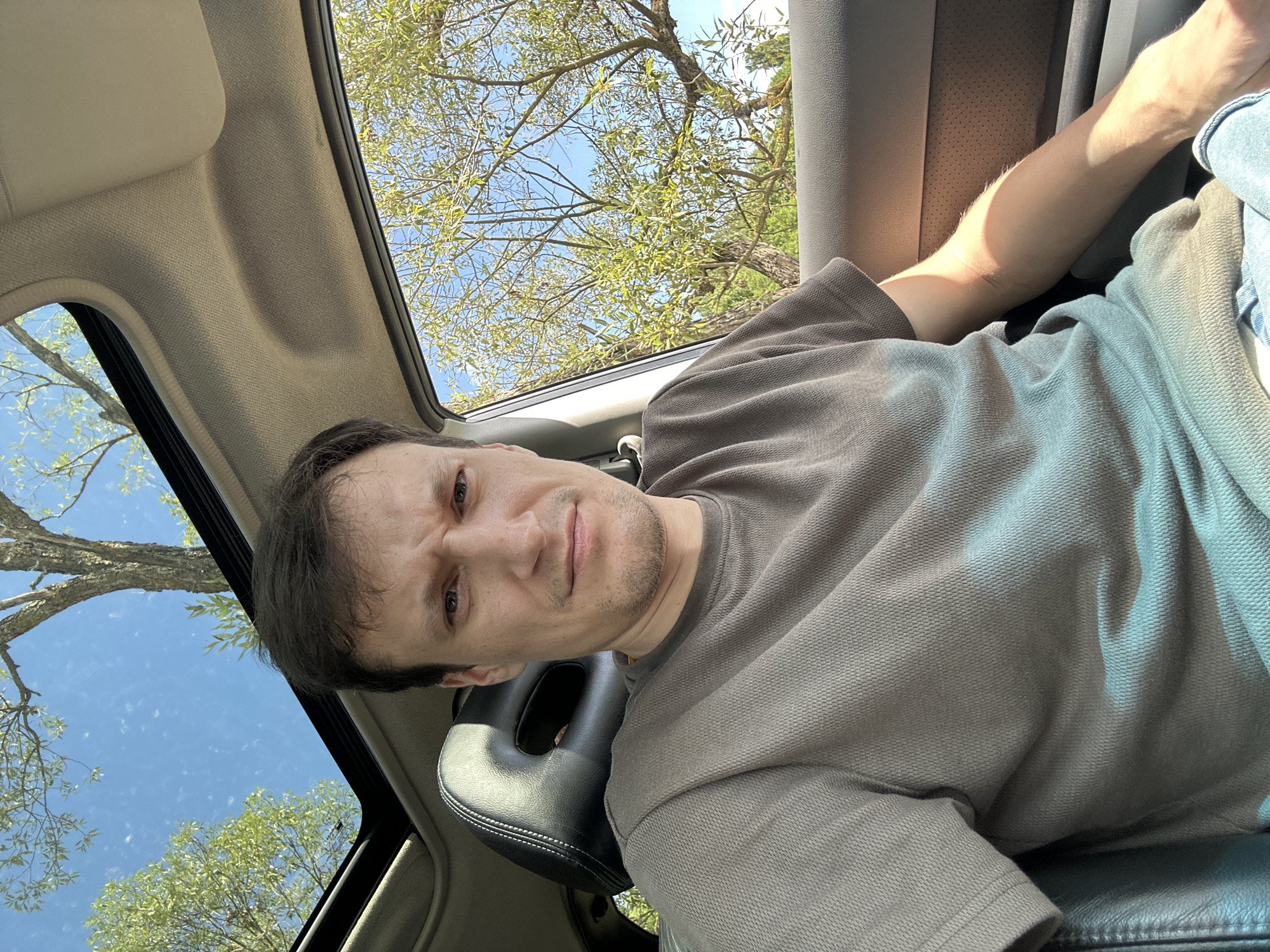 A man with short brown hair and light skin is sitting in the driver's seat of a car, looking directly at the camera with a slight frown. He is wearing a short-sleeved, textured grey t-shirt and what appear to be blue jeans. The car has a black leather headrest visible behind him, and the interior is a neutral tan color. Through the open sunroof and the passenger-side window, lush green trees and a bright blue sky are visible, suggesting it is daytime and likely a warm day. The overall impression is a casual, candid shot taken from inside a vehicle, possibly in a rural or suburban setting given the prominent natural surroundings. The specific location in Makiv, Ukraine, cannot be determined from the visual information provided.