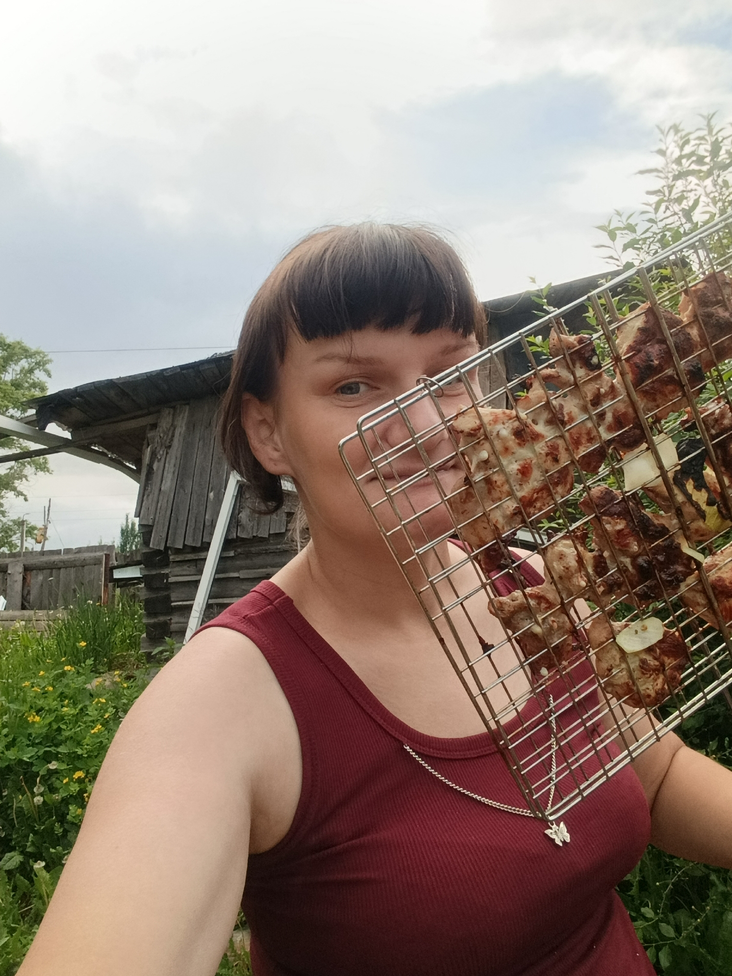 An outdoor, eye-level selfie-style photograph depicts a Caucasian female subject in the city of Фоки, Russia, holding a metal barbecue grill rack laden with cooked meat and onion slices directly in front of her face. The woman has short, dark brown hair with bangs and is wearing a sleeveless, ribbed, maroon tank top with a silver chain necklace featuring a butterfly pendant. Her left eye is visible, looking directly at the camera with a slight smile, while the grill obscures most of her nose and mouth. The grill contains multiple pieces of browned, possibly charred, meat along with white onion slices. The background consists of an overcast sky, dense green vegetation with yellow wildflowers, and a weathered wooden structure with a dark roof and vertical planks situated behind her left shoulder. A partial wooden fence is also visible in the mid-ground.