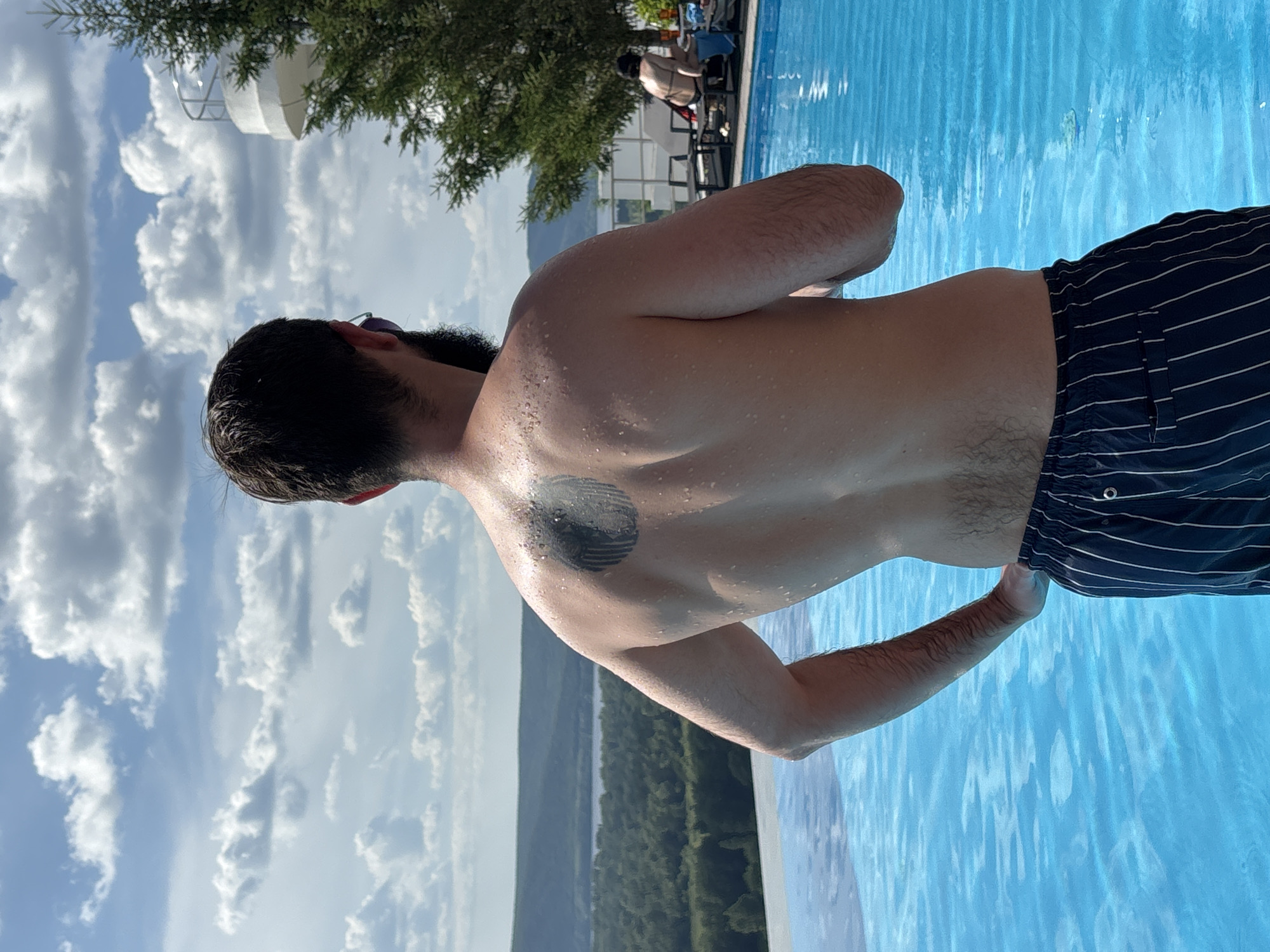 The image depicts a man standing at the edge of an infinity pool, with his back to the camera. He has a dark beard and is wearing dark blue striped swim trunks. A dark circular tattoo is visible on his upper back, and his skin is speckled with water droplets. The pool's bright blue water reflects the sky, which is filled with scattered white clouds under a bright sky. In the background, a lush green forest stretches across rolling hills, with a body of water visible in the distance. To the right of the man, a large evergreen tree stands near a white, multi-level structure, possibly a building or observation deck. Further in the background, other people are visible relaxing on lounge chairs near the pool. The overall scene suggests a summer day at a resort or scenic overlook, likely in the outskirts of Novokuybyshevsk, Russia, given the natural landscape.