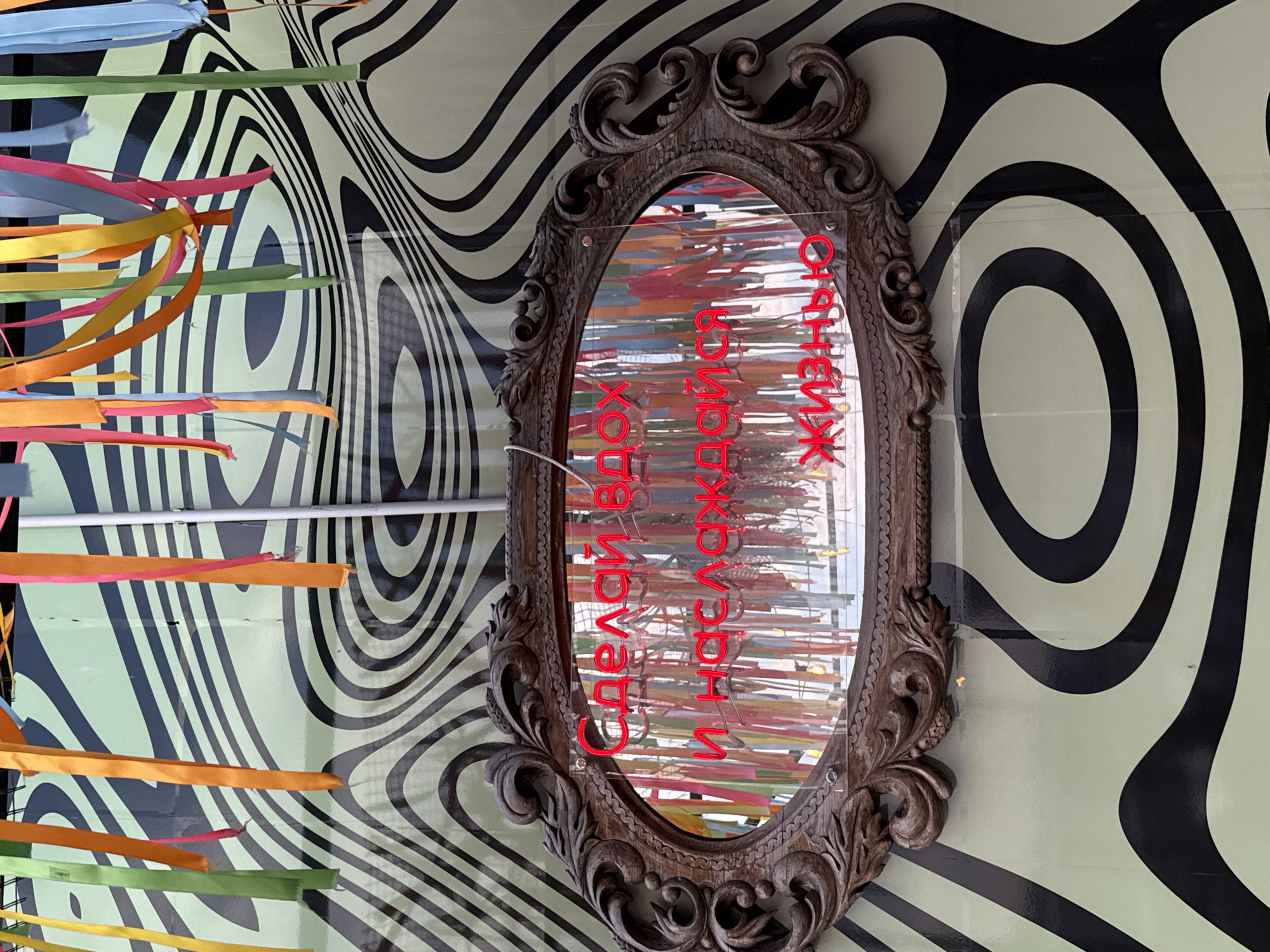 In Izhevsk, Russia, a vintage-style, ornate wooden mirror is mounted on a wall with a psychedelic, black and lime green wavy pattern. Colorful ribbons hang vertically from the top of the wall, some passing behind the mirror. Red neon text is visible within the mirror's reflection, reading "Сделай вдох и наслаждайся жизнью" which translates to "Take a breath and enjoy life." The scene appears to be an outdoor or semi-outdoor public space, possibly a decorative wall or installation.