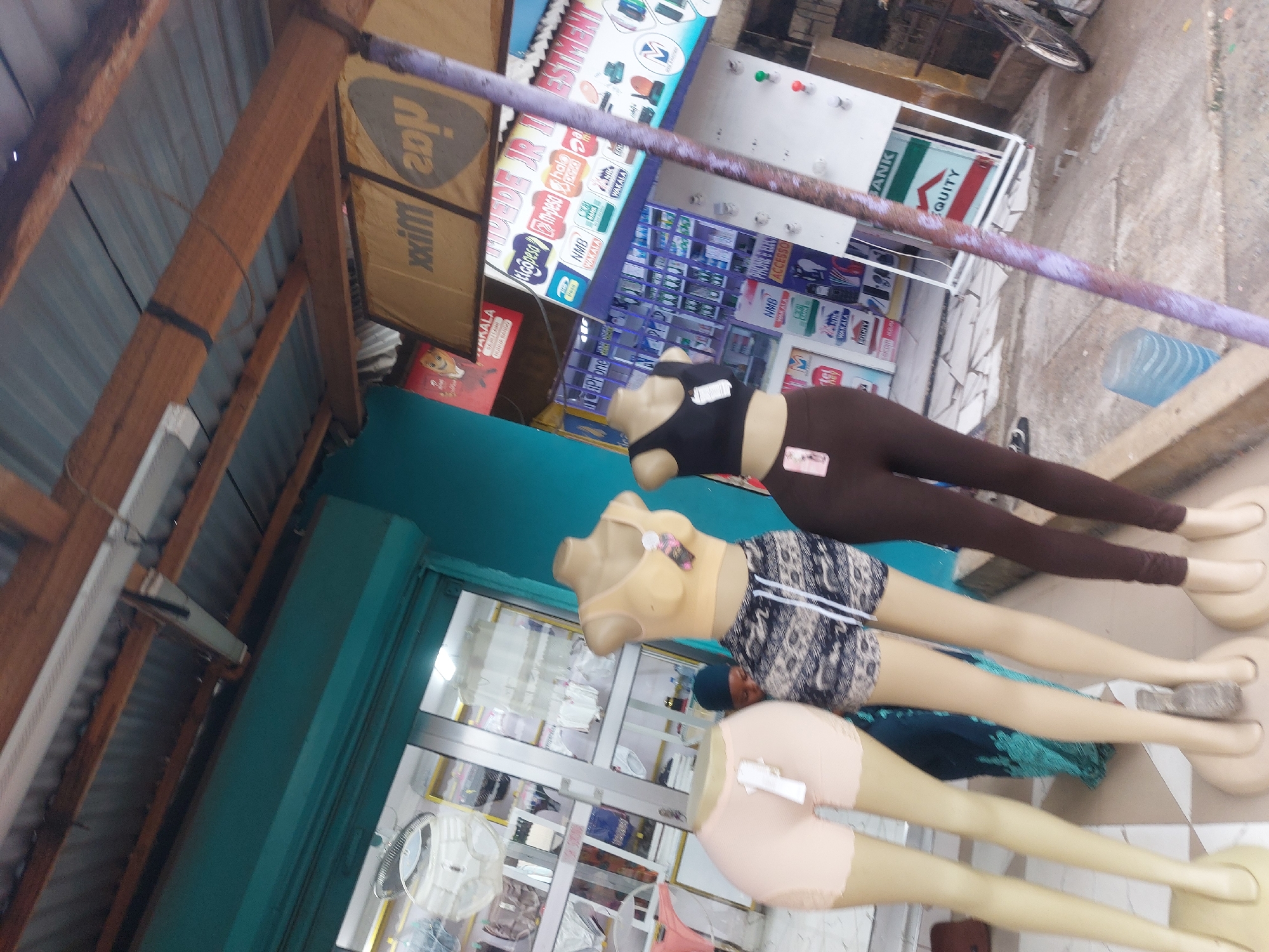 Here's a detailed description of the image:
**Setting:** A street scene, likely in a commercial area. There are structures resembling storefronts or businesses.
**Objects:**
* **Mannequins:** Three female mannequins. They are dressed in clothing and are posed horizontally.
* **Advertisements and Signage:** There are numerous signs and advertisements visible in the background. Brands like "NMB", "Tigo", "M-Pesa" are recognizable.
* **Building Structure:** Portions of a building are visible, including a roof structure with wooden beams and metal sheeting.
* **Assortment of items inside a glass door:** Clothing and textiles seem to be on display behind the mannequins.
* **Pipe or Rail:** There is a pipe or rail running horizontally across the image.
**Environment:**
* **Urban/Commercial:** The scene suggests a busy urban environment with commercial activities.
**Location Clues:**
* **Tanzania:** The presence of Swahili words (such as "Wakala" and "Mpesa") on the advertisements indicates the location is in Tanzania.
* **Dar es Salaam:** The brands like NMB, Tigo, and M-Pesa are commonly found in Dar es Salaam.