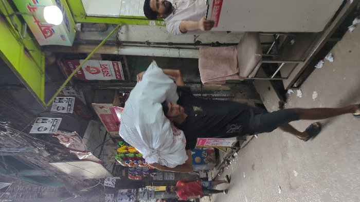 The image depicts a narrow, bustling commercial street or alleyway within an urban area of Dhaka, Bangladesh, under daylight conditions.
In the foreground, a young adult male, likely in his late teens or early twenties, with dark hair and a light beard, walks from right to left. He is wearing a black short-sleeved t-shirt with a white design, dark knee-length shorts, and black slide sandals. He carries a large, white, irregularly shaped sack on his left shoulder and head, supporting it with both hands.
In the mid-ground on the left, a second young male, wearing a red t-shirt and light denim shorts, walks away from the camera. On the right, a third male, with a beard and wearing a light-colored polo shirt, is partially visible, positioned at what appears to be a shop entrance, with his right hand resting on a white counter or appliance.
The street surface is paved and cluttered with scattered white litter, including crumpled papers and plastic. Buildings rise on both sides of the alley. On the right, a shop entrance features a prominent lime-green awning, a spherical white light fixture, and signs with Bengali script, notably the "Nagad" logo. A light-colored upholstered chair with a metal frame and a towel draped over it is placed on a raised platform outside this shop. On the left, another shopfront displays colorful packaged goods hanging for sale, and numerous political or advertising posters featuring faces and Bengali text are affixed to the building walls. A dense network of black electrical or communication cables crisscrosses overhead between the buildings.