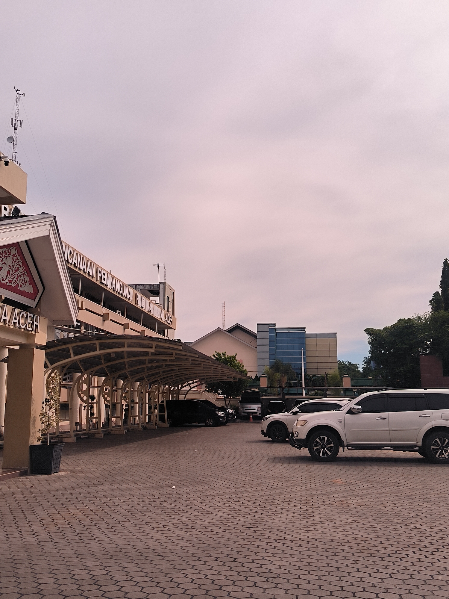 This is a vertical, eye-level shot of a paved parking area in front of a building complex in Banda Aceh, Indonesia. The sky is overcast with a soft, diffused light, suggesting it is either early morning or late afternoon. The scene is dominated by the architecture of the buildings and the parked vehicles.
In the foreground, the paving stones are laid in a hexagonal pattern, reflecting the muted light. To the left, a building entrance is partially visible, featuring a prominent sign with text that appears to be in Indonesian. A covered walkway with a curved metal roof extends from this building, supported by pillars, and provides shade over a portion of the parking area. A black SUV is parked under this walkway.
Further into the scene, several white SUVs are parked in the open. One is prominently in the mid-ground, facing left, its wheels turned. Beyond the SUVs, more buildings are visible, including a modern structure with blue glass windows and a lighter-colored building with several antennas on its roof, indicating a communication tower. Lush green trees are present to the right, bordering the complex.
The overall mood is calm and somewhat serene, with the overcast sky creating a soft, even light. There is no visible human activity, and the focus is on the structures and vehicles in the parking lot. The visible text on the building includes "NCAAAN PEMBANGUNA RIA ACEH" and "DA ACEH".