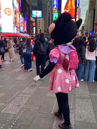 The image shows a person dressed in a Minnie Mouse costume in Times Square, New York City. The costumed figure is wearing a pink dress with white polka dots, black leggings, and brown shoes. They also have a small pink Nike backpack. The background consists of a crowded street with pedestrians, tall buildings, and digital billboards. The ground is paved with grey stone tiles.