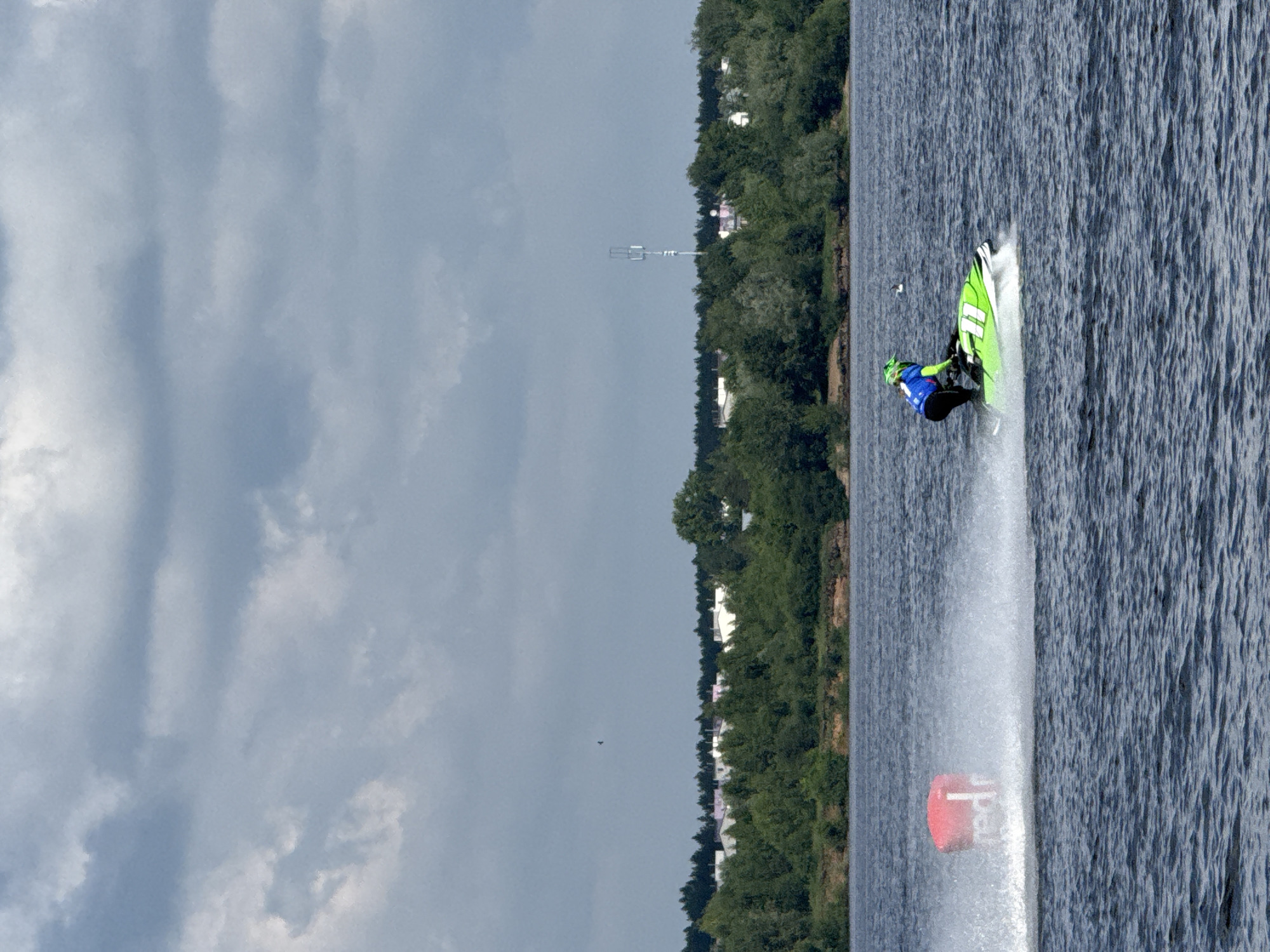 This is a photograph taken from a distance, showing a jet ski rider on a body of water, likely a river. The rider is wearing a blue and green racing suit and a helmet. They are on a green and white jet ski, which is kicking up a significant amount of spray as it moves. In the background, there is a shoreline with a dense line of green trees and some white buildings visible behind them. Further in the distance, a cell tower can be seen. The sky is overcast with dramatic, dark grey clouds. To the left of the jet ski, a red buoy with white lettering, appearing to say "red", is visible in the water, also creating spray. The water itself is dark and appears choppy. The general impression is of a water sport event taking place on a cloudy day. The presence of the cell tower and the style of the buildings suggest an urban or suburban environment. Given the mention of the city of Perm, Russia, it is probable that this scene is depicting a jet ski race or recreational activity on one of the city's waterways, such as the Kama River.