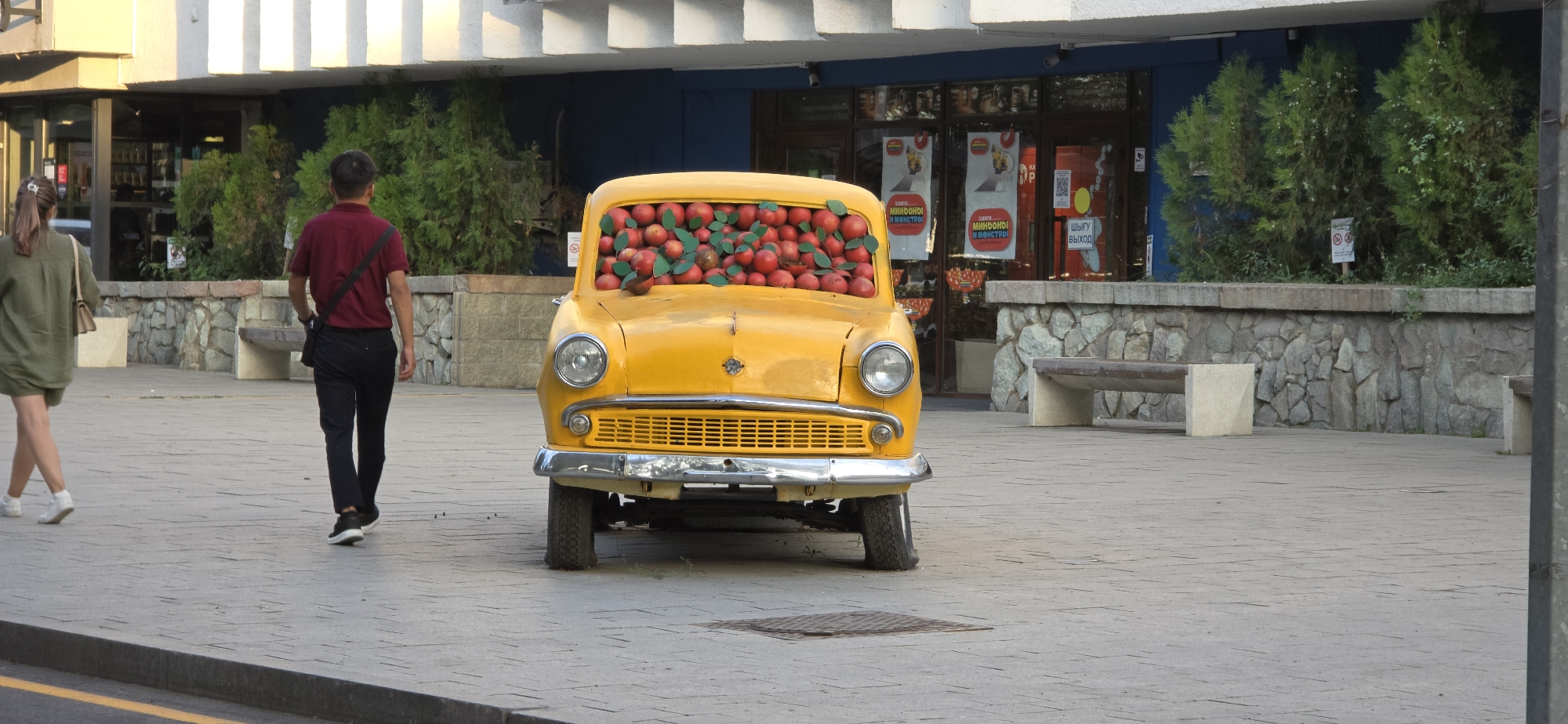 The image depicts a street scene in Almaty, Kazakhstan, featuring a bright yellow vintage car parked on a paved pedestrian walkway. The car's front windshield is filled with red apples and green leaves, suggesting a decorative display. Two people are walking on the walkway. On the left, a woman with long brown hair tied in a ponytail wears a green dress and carries a small purse. On the right, a man with short dark hair walks away from the camera, wearing a maroon polo shirt and dark pants, with a black bag slung across his body. Behind the car, a building with large glass windows is visible, along with several green bushes and trees. Stone walls and benches are also part of the urban landscape. The overall lighting suggests it is daytime, with the sun casting soft shadows.