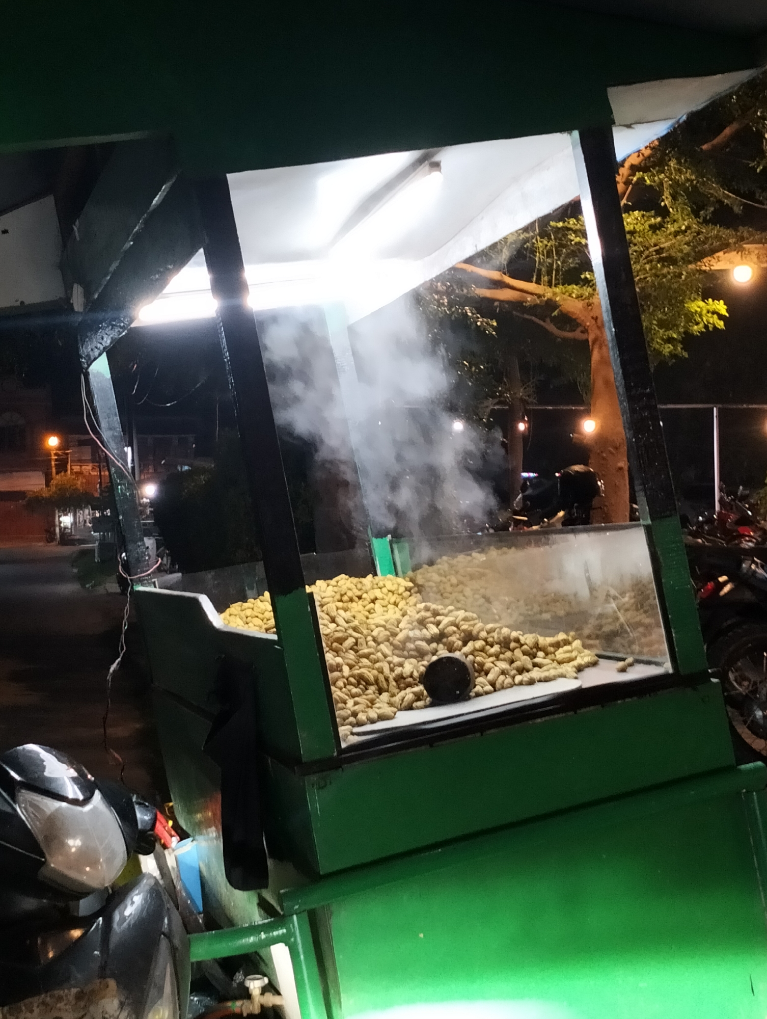 A food cart full of steaming peanuts is positioned on a street in Lhokseumawe, Indonesia, at night. The cart is painted green and has a white light above it. A plume of steam rises from the peanuts inside a clear display case. A parked motorbike is partially visible in the foreground. Other parked motorbikes are behind the cart. Streetlights and building lights provide illumination in the background. A tree with sparse foliage is to the right of the cart. The street appears wet.