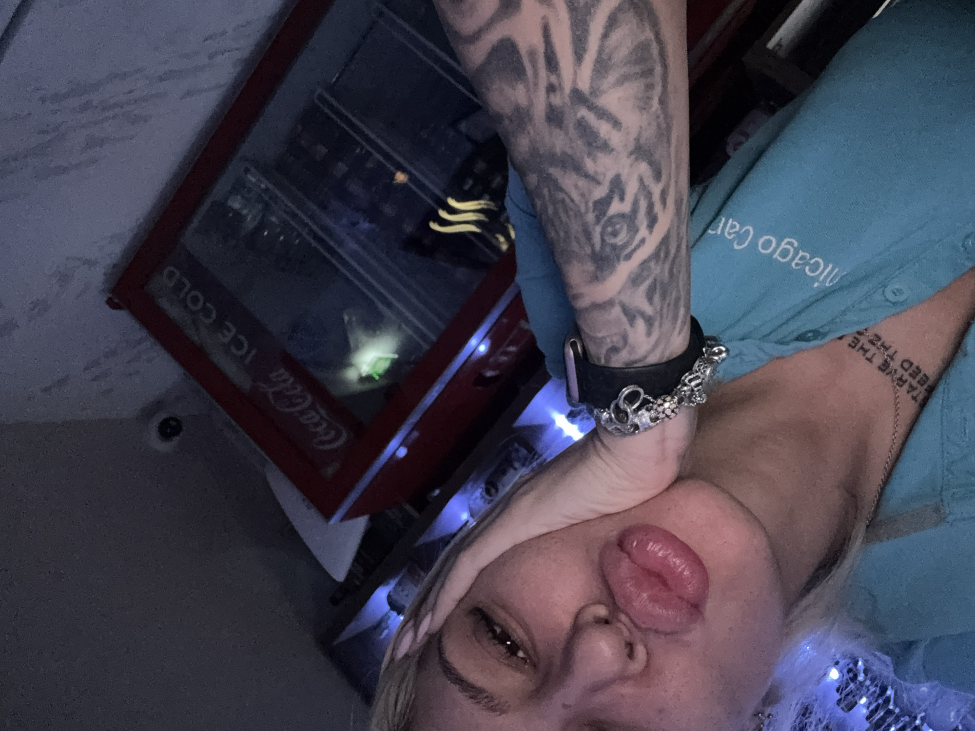 A person with blonde hair is posing indoors near a Coca-Cola refrigerator. The person has a tattoo of a wolf's head on their forearm and a bracelet on their wrist. They are wearing a light blue shirt with the words "Chicago Car" written on it. The location is Plovdiv, Bulgaria.