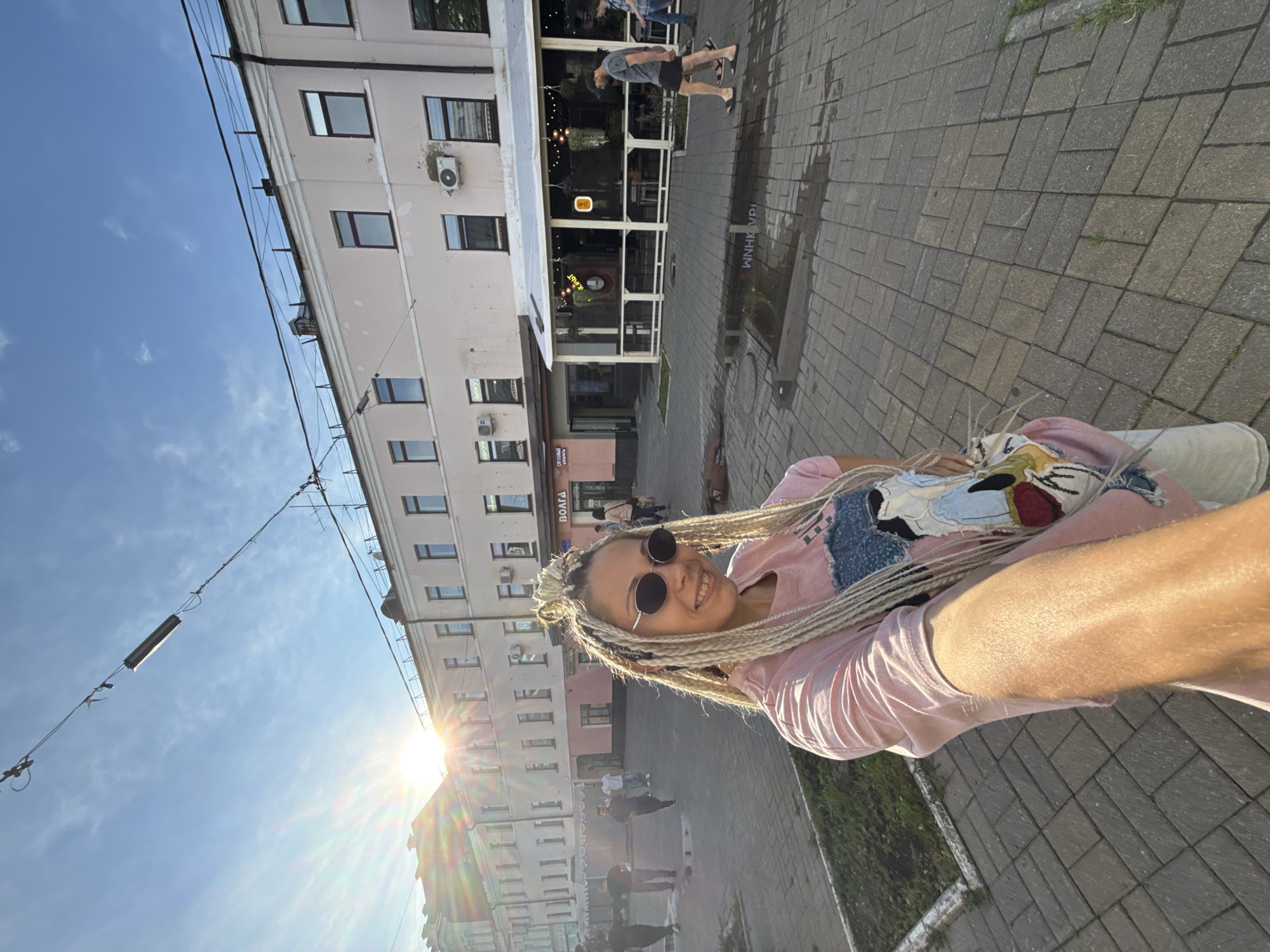A young woman with blonde braided hair and sunglasses is taking a selfie in a city square in Yaroslavl', Russia. She is wearing a pink t-shirt with a colorful graphic print and white shorts. She is standing on a paved area with a grassy strip to her left. In the background, there is a light pink building with multiple windows and an outdoor seating area for a cafe. The sun is shining brightly, creating lens flare and casting long shadows. Several other people are walking in the square. The overall atmosphere is bright and casual.