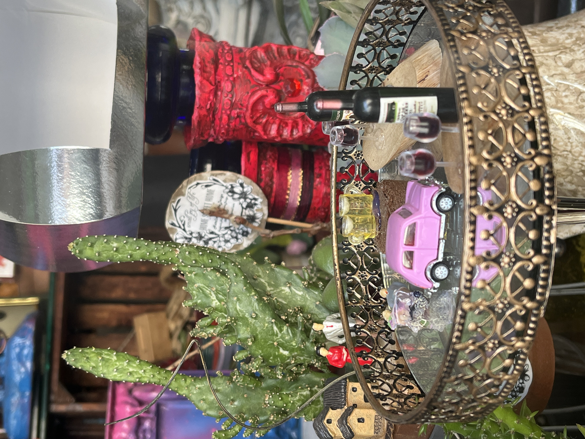 The image shows a still life with various objects, including a cactus, a red ornate object, a decorative metal hoop, a small wine bottle, miniature wine glasses, two pink toy cars, a label with floral designs, and other assorted items. The arrangement is indoors. There is no verifiable indication this image was taken in Varna, Bulgaria.
