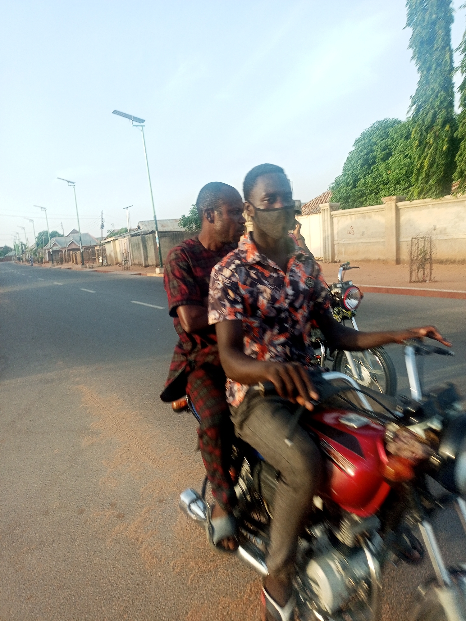 Two men ride a red motorcycle down a paved road in Tempure, Nigeria. The driver wears a patterned shirt, grey trousers, and a face mask. The passenger behind him is in a red and black plaid shirt and trousers. Both are wearing sandals. In the background is another motorcycle, streetlights, buildings, and trees.