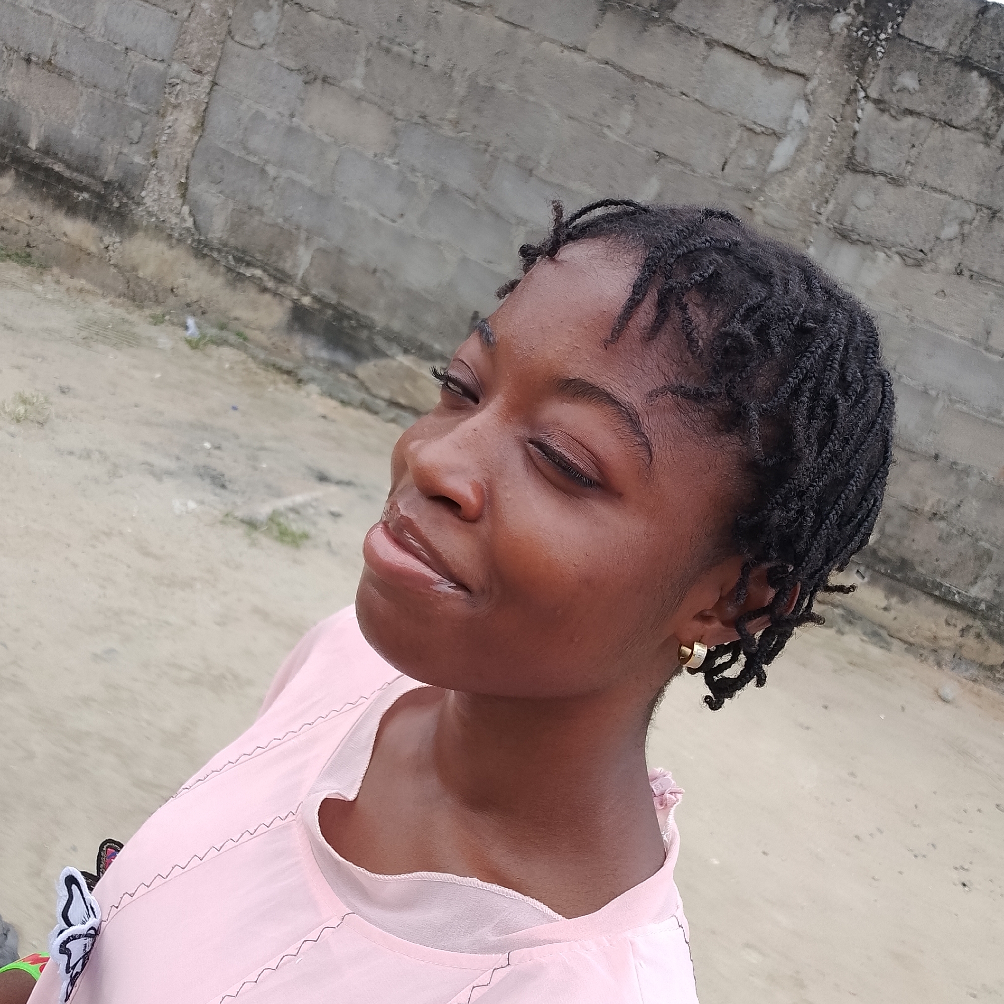 In Ado, Nigeria, a young Black woman with dark braided hair and gold hoop earrings is captured in a medium, close-up portrait. She is wearing a light pink top with a textured neckline. Her head is tilted back slightly, and her eyes are closed as she looks upwards, with her lips parted. The background consists of a textured concrete wall and a sandy, somewhat barren ground. The lighting suggests an outdoor setting.