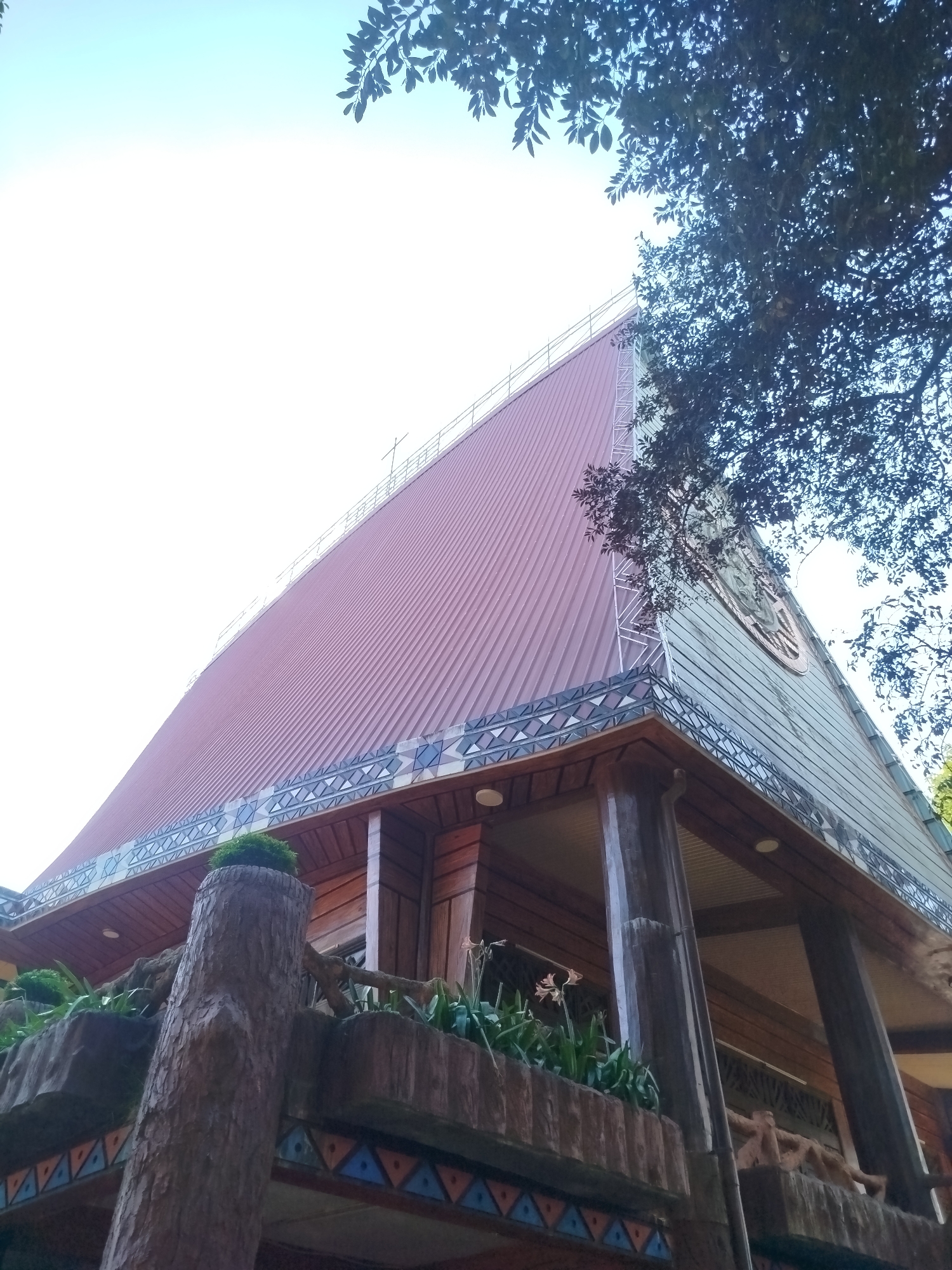 A low-angle shot of a building, possibly a church, located in An Phu Ward, Vietnam. The building features a high-pitched roof made of brown corrugated metal, decorated with a border of repeating geometric patterns. A cross is located at the top of the roofline. The building's facade includes a carved emblem and is supported by dark, cylindrical pillars. A decorative balcony with plant life and faux-wood pillars stands in the foreground. The sky is bright, with foliage from nearby trees framing the top of the image.