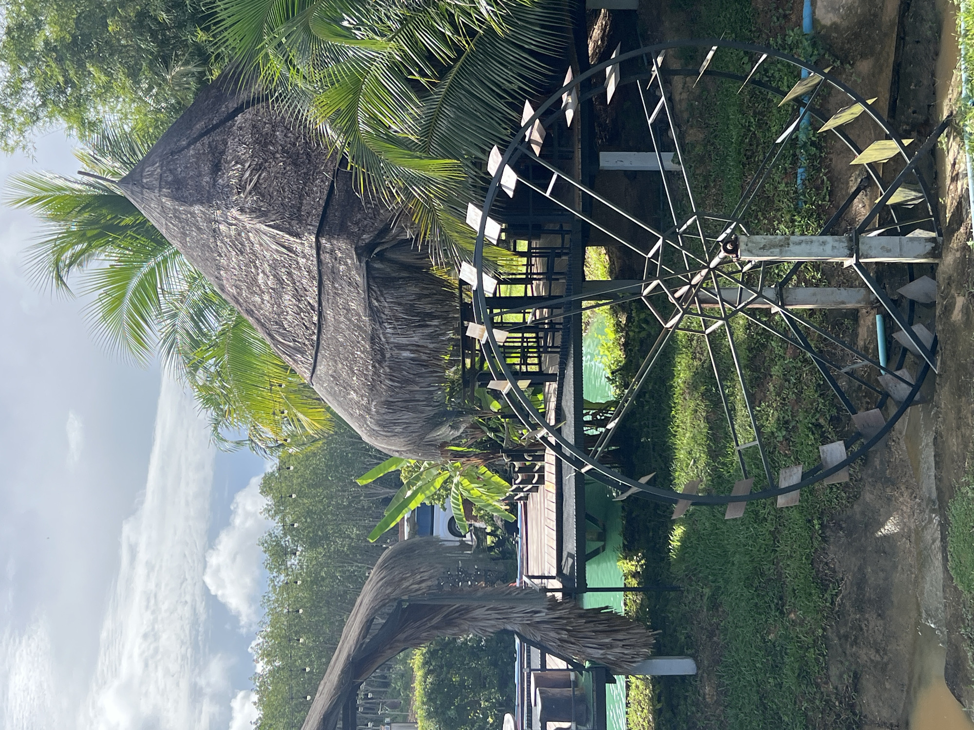 This image depicts a scenic outdoor scene in Tambon Pa Kae Bo Hin, Thailand. A prominent feature is a large, metal water wheel positioned in the foreground, with its spokes and paddles visible. Behind the water wheel, a wooden deck is elevated over a body of turquoise water. On the deck, there are several thatched-roof structures, one of which is conical and appears to be a hut or pavilion. Palm trees and lush green vegetation surround the area, contributing to the tropical ambiance. The sky above is partly cloudy, with a mix of white clouds and blue sky. In the background, a line of trees can be seen, and further in the distance, a white vehicle is partially visible. The ground around the water wheel is covered in grass and soil, with some areas of exposed earth. The overall impression is one of a tranquil, nature-infused setting, likely a resort or recreational area.