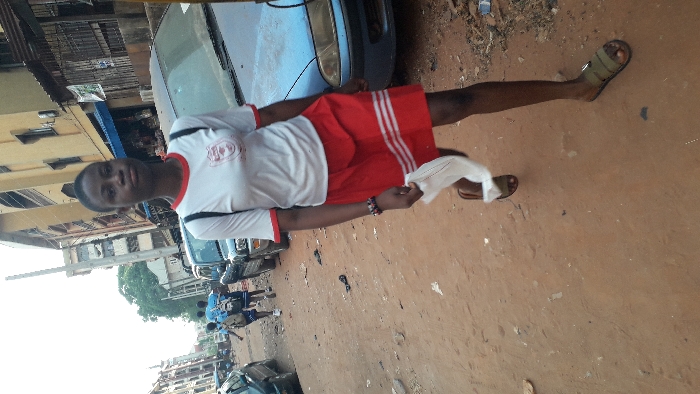 A young female, of dark complexion, walks on an unpaved, reddish-brown dirt street in Onitsha, Nigeria. She is positioned centrally, moving forward while looking towards the camera with a neutral expression. She wears a white short-sleeved t-shirt with red collar and cuffs, featuring a red-and-white crest on the left chest, and a red skirt with three horizontal white stripes near the hem. A black backpack is on her shoulders, a multi-colored bracelet on her right wrist, and olive-green slide sandals on her feet. She holds a crumpled white object in her left hand.
Immediately to her right is a parked, faded blue sedan. In the mid-ground, further down the street, several individuals, appearing to be students in blue shirts and carrying backpacks, walk away from the camera. The street is lined on both sides by multi-story concrete buildings, some with visible balconies and ground-floor shopfronts, including a prominent yellow/beige building on the right. Additional parked vehicles are visible along the street. A tall, light-colored utility pole stands in the mid-ground. The street surface is earthen with scattered debris. The sky above is bright and overcast.