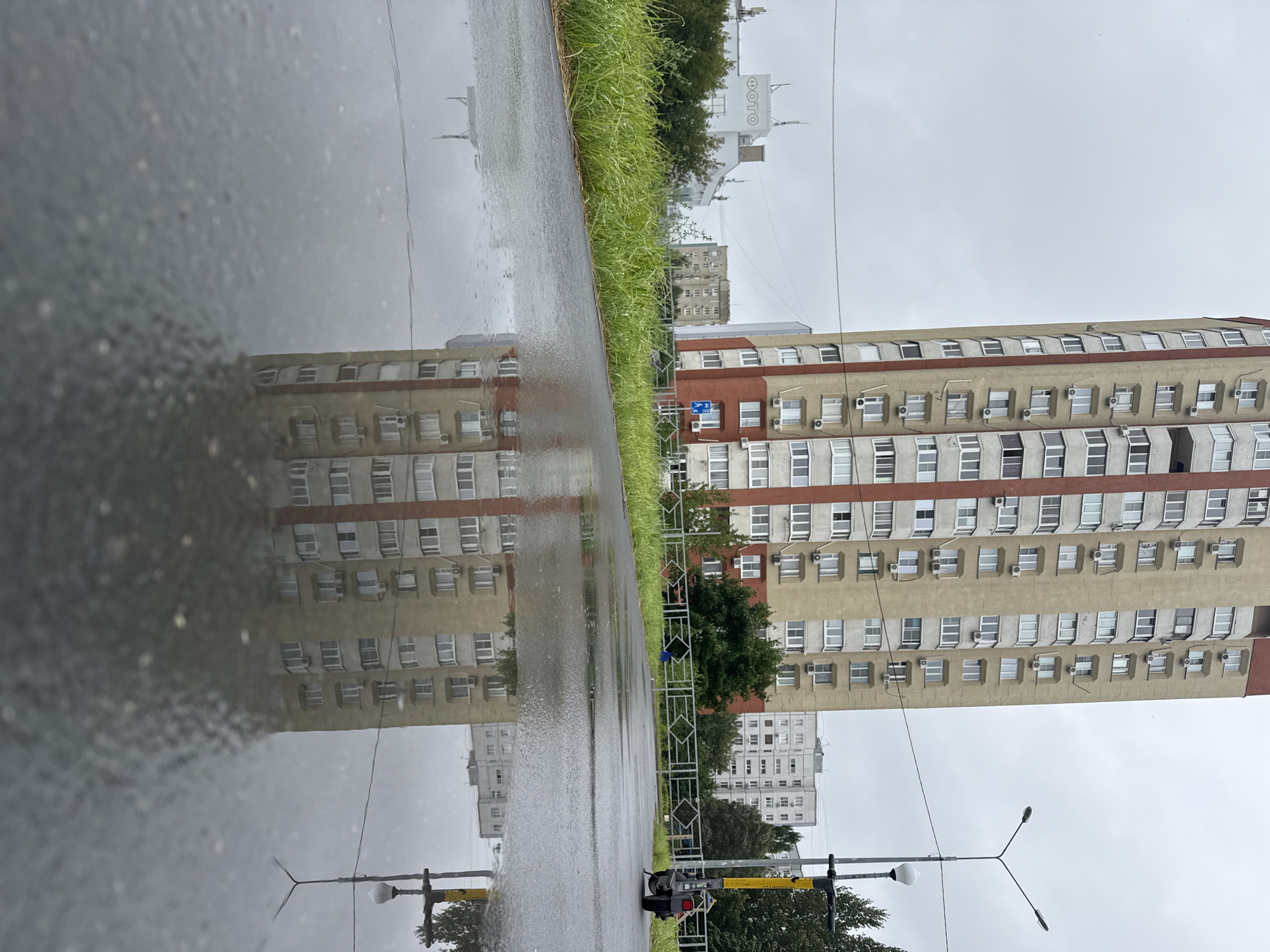 The image depicts a rainy day in Tolyatti, Russia, specifically in an urban environment with residential buildings and a paved area. In the foreground, a puddle reflects a tall, multi-story apartment building. The building features a beige facade with vertical red accents and numerous windows, many with air conditioning units installed. The reflection in the puddle is clear, showing the architectural details of the building and its multiple stories.
Behind the puddle, a paved path or road is visible, bordered by a patch of green grass on the left. Beyond the grass, another, shorter apartment building can be seen, with the word "ФОТО" (which means "PHOTO" in Russian) in large white letters on its facade. Further in the background, more residential buildings and trees are present under an overcast sky.
To the right of the scene, a modern street light with a dual lamp head stands tall, and next to it, a yellow and black electric scooter is parked. The scooter has a distinctive yellow stem and a black body, with its rear wheel visible. A metal fence with a geometric pattern separates the paved area from the grass and the buildings. The overall atmosphere is one of a subdued, post-rain urban landscape.