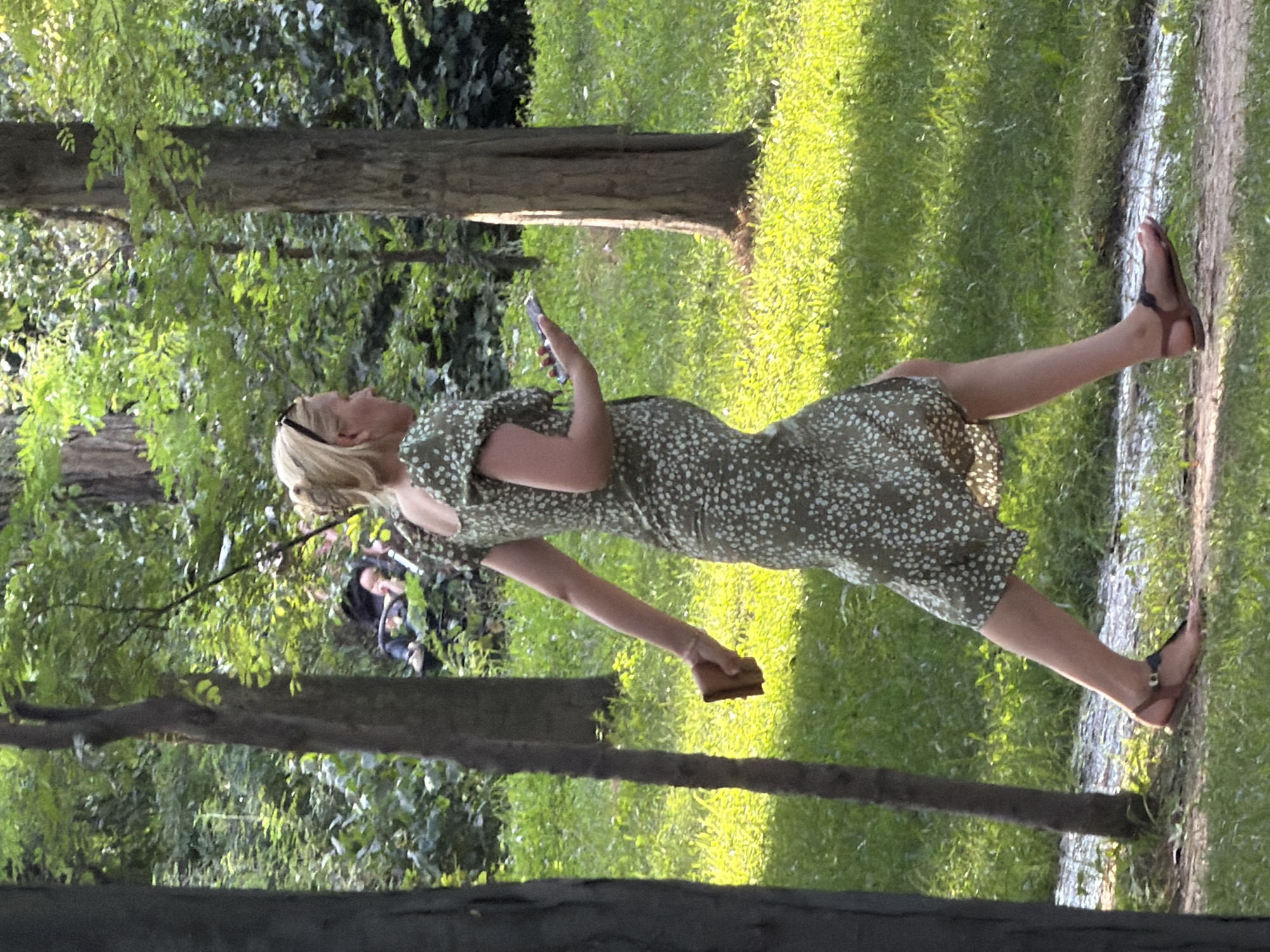 A blonde woman in a green floral dress and brown sandals walks on a path in a park. She is holding a cell phone in her right hand and a small brown object, possibly a piece of food, in her left. In the background, two people are lying down on the grass, partially obscured by trees and foliage. The scene is bathed in dappled sunlight.