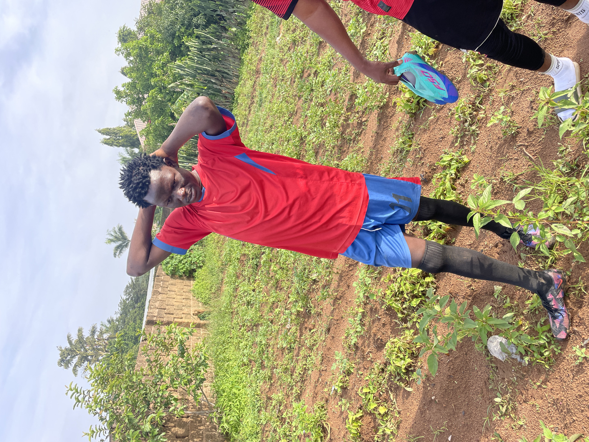 In Gura Top, Nigeria, a young man stands in a field wearing a red and blue soccer jersey, blue shorts, and black socks. His hands are clasped behind his head. To his right, another person, partially visible, is holding a turquoise and purple soccer cleat. The field is covered in dirt and small green plants, with a brick wall and trees in the background under a cloudy sky.
