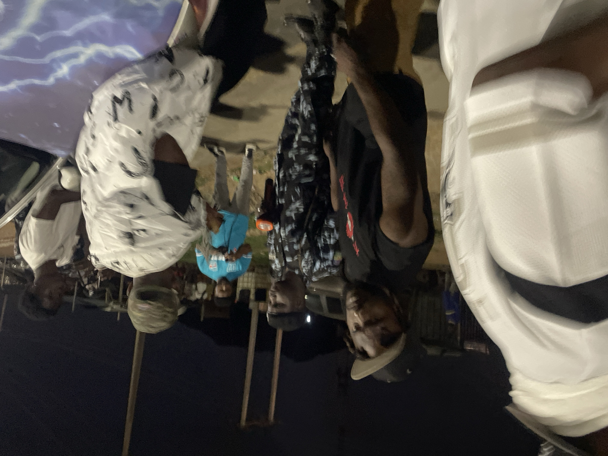 The image shows a group of people, potentially a street gathering, in what is presumed to be Kubwa, Nigeria, given the prompt context. The lighting suggests it's nighttime.
People: Several individuals are visible, wearing various clothing styles. One is wearing a white and black patterned shirt, another a blue t-shirt, and at least one is in a black shirt. Two individuals appear to be wearing what looks like a Police uniform.
Objects: There are some poles visible. In the background, there appear to be automobiles.
Environment: The scene is at night. The ground is paved, possibly a street or open area.