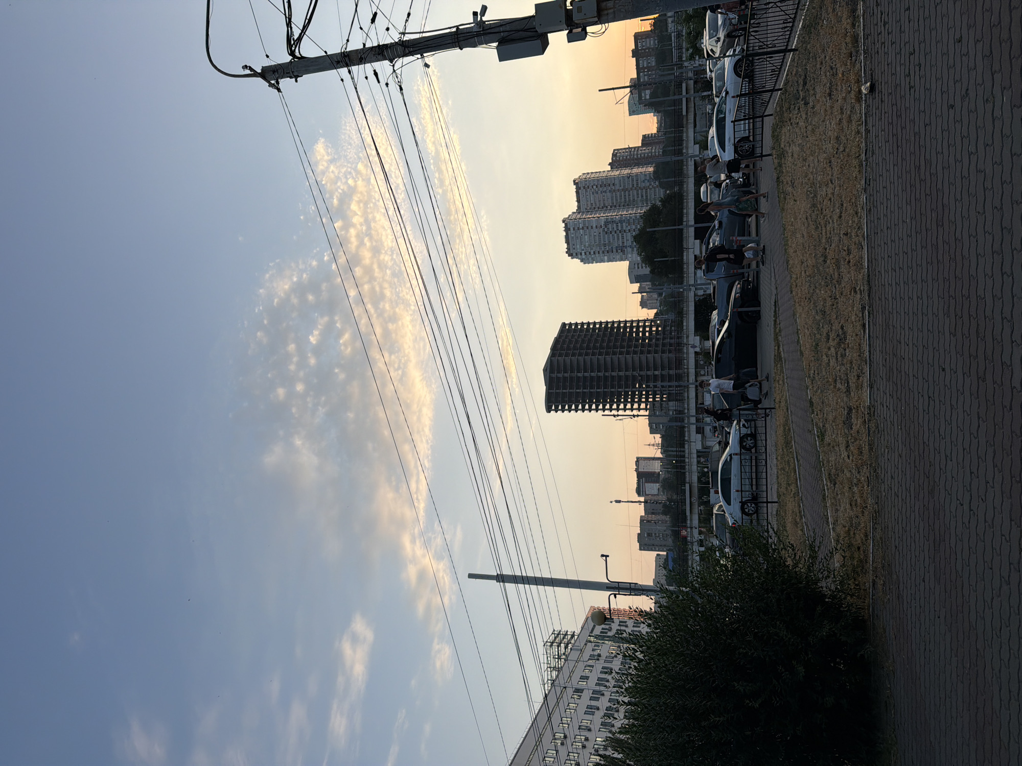 The image shows a street scene in Rostov-on-Don, Russia, during a sunset. The sky is a mix of blue and orange hues, with scattered clouds. In the foreground, a paved walkway is visible, leading to a grassy area with dry vegetation. A black metal fence separates the walkway from a parking lot where several cars are parked.
Several people are present in the scene, some walking and others standing. They are dressed in casual clothing, suggesting a relaxed atmosphere. In the background, tall buildings and construction sites dominate the skyline, indicating an urban environment. A utility pole with numerous wires is also prominently featured on the right side of the image. The overall impression is that of a typical evening in a Russian city, with a blend of urban infrastructure and natural elements.