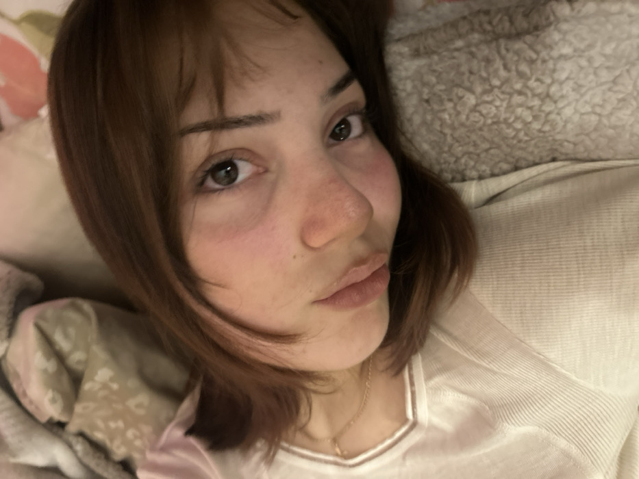A close-up, slightly angled selfie captures a woman with light skin and shoulder-length brown hair. Her expression is soft, with a gentle pursing of her lips. She wears a light-colored, possibly white or cream, top with a simple neckline. A delicate gold necklace is visible. The background includes soft, textured bedding, suggesting a comfortable and relaxed setting. Her gaze is directed toward the camera, with dark eyes that appear tired. The lighting is soft, highlighting the contours of her face.