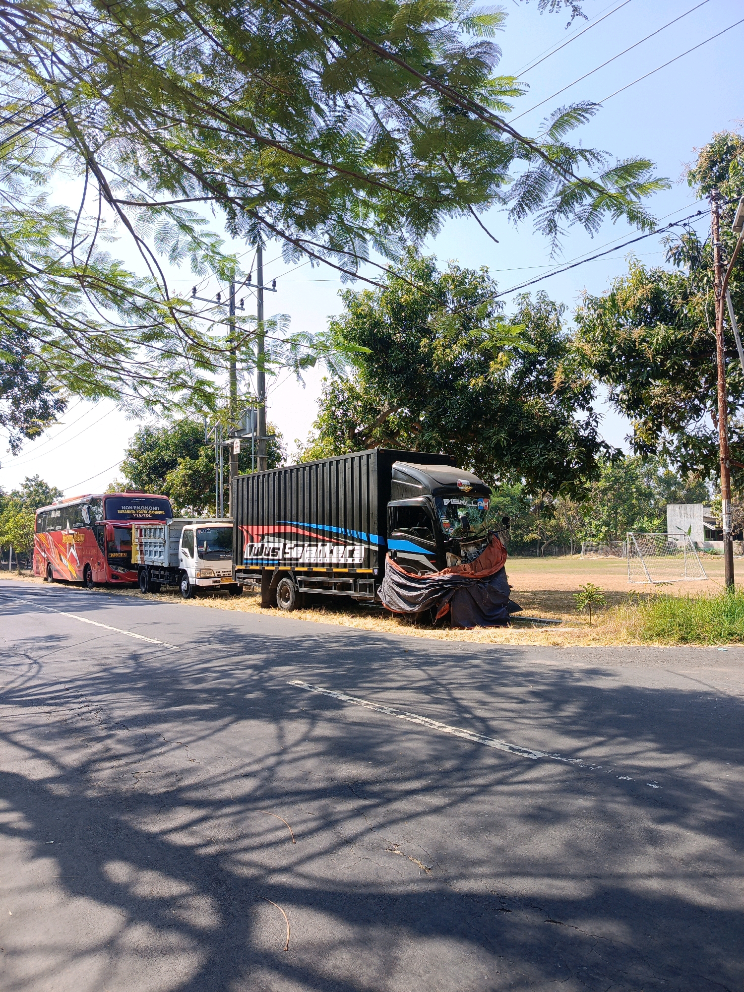 The image depicts a roadside scene in Tambran, Indonesia. In the foreground, a paved road with white lane markings is visible, with dappled shadows of tree branches cast across its surface. On the side of the road, several vehicles are parked. Closest to the foreground is a large black truck with a ribbed container. Its front is partially covered with a draped dark cloth. To its left is a smaller white truck. Behind the white truck is a red bus with yellow and white decals. The background shows a grassy field with a white structure and a soccer goal, indicating a sports field. Trees line the edge of the field, and utility poles with wires are present. The sky is clear and blue. The scene appears to be during the daytime with bright sunlight. No people are visible in the image.