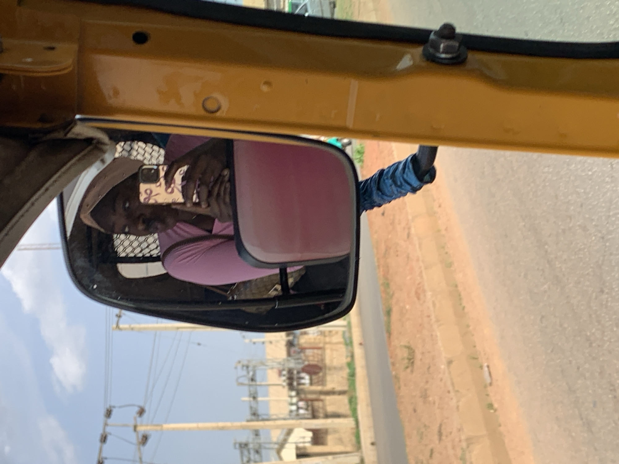 A woman in a pink shirt and light-colored hat is reflected in the side mirror of a yellow vehicle. The woman is holding a cell phone with a patterned case. Outside the vehicle, there is a street with power lines and utility infrastructure. The location is Rafin Par, Nigeria. The ground appears to be a mixture of dirt and pavement.