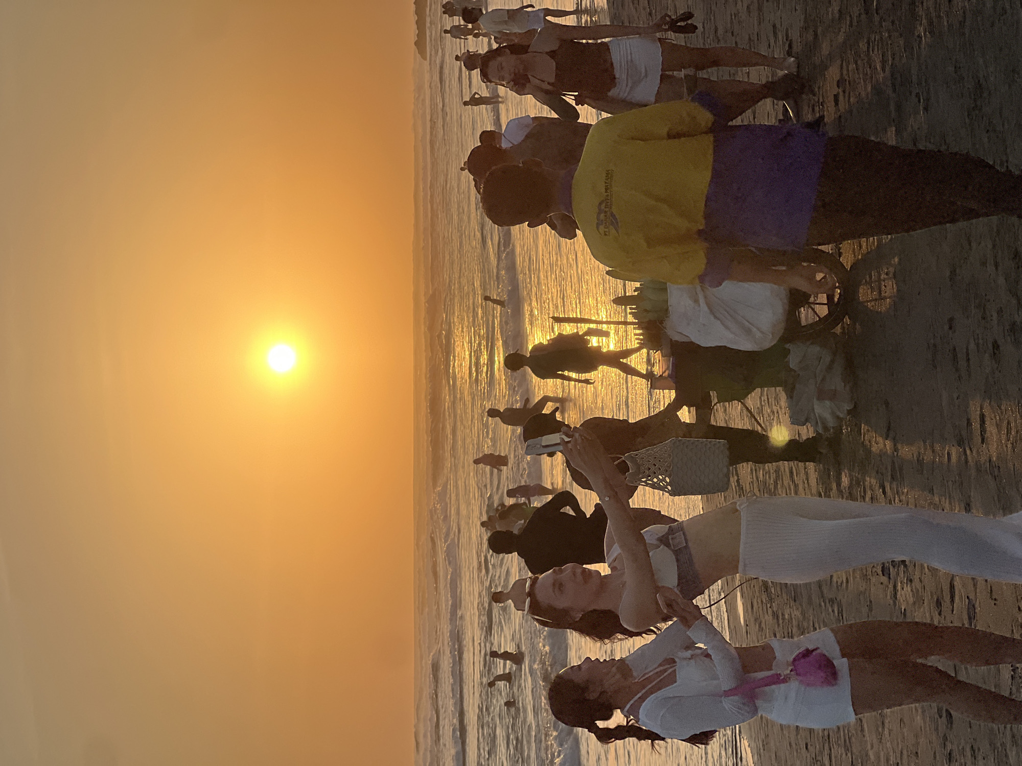 The image depicts a crowded beach scene at sunset in North Kuta, Indonesia. The sky is illuminated by a vibrant orange glow as the sun sets over the ocean, casting a warm light on the water and the sand. Numerous people are visible in the shallow water, wading and enjoying the waves. On the shore, several individuals are present, including two women in the foreground taking a selfie. One woman on the left wears a white top and shorts with a pink handbag, while the woman on the right is dressed in a white top and pants. Behind them, a vendor with a cart filled with corn is positioned near other beachgoers. The overall atmosphere is lively, with people engaged in various activities such as swimming, walking, and socializing.