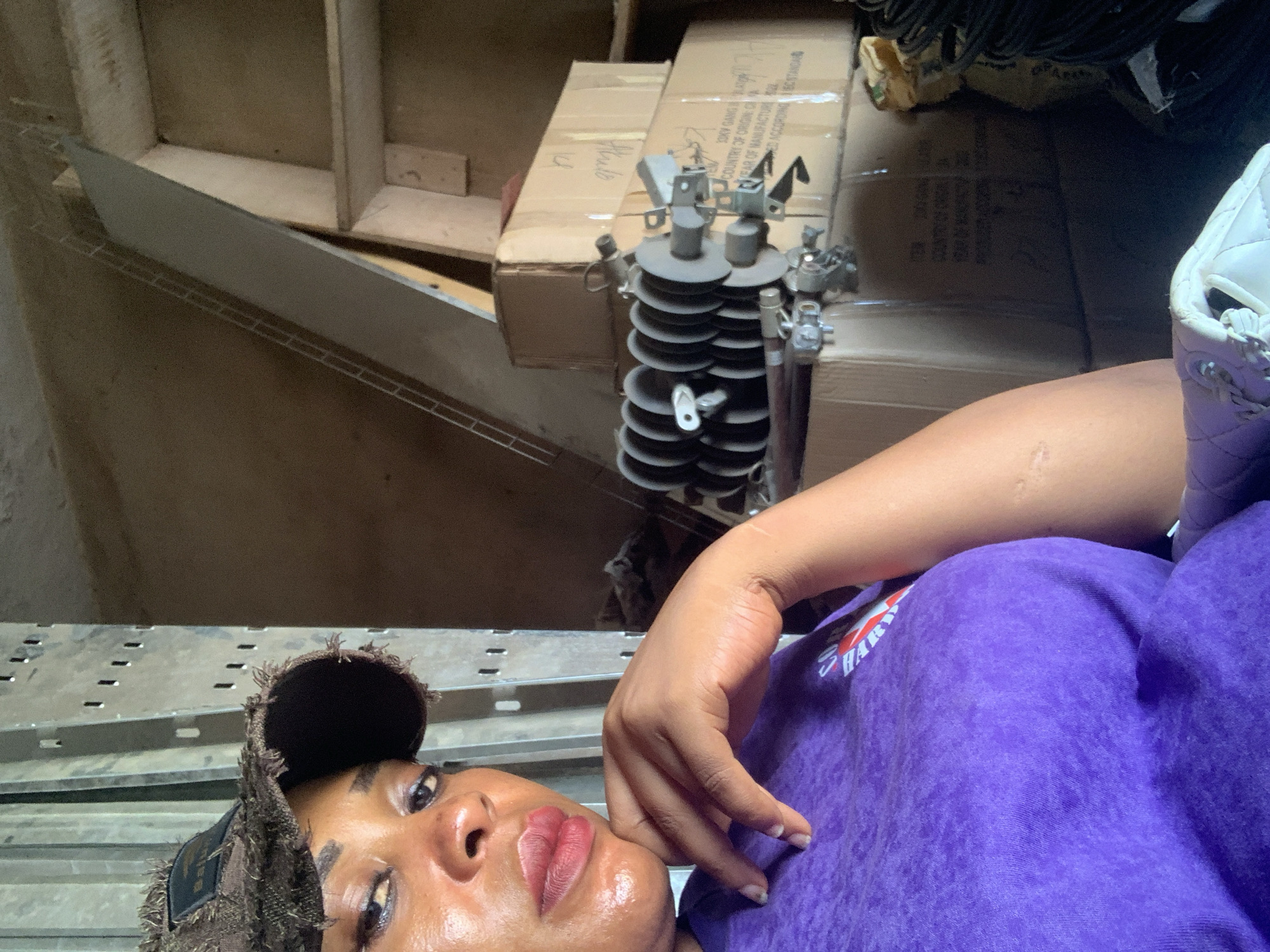 A close-up, high-angle shot depicts a person with dark skin lying down, partially visible. Their face, with glossy lips, is framed by a fringed cap. They are wearing a purple sweatshirt with visible lettering. Behind them are boxes and what appears to be electrical equipment. The setting seems to be an interior space with shelving and various objects stored, which is located in the city of Shaba, Nigeria.